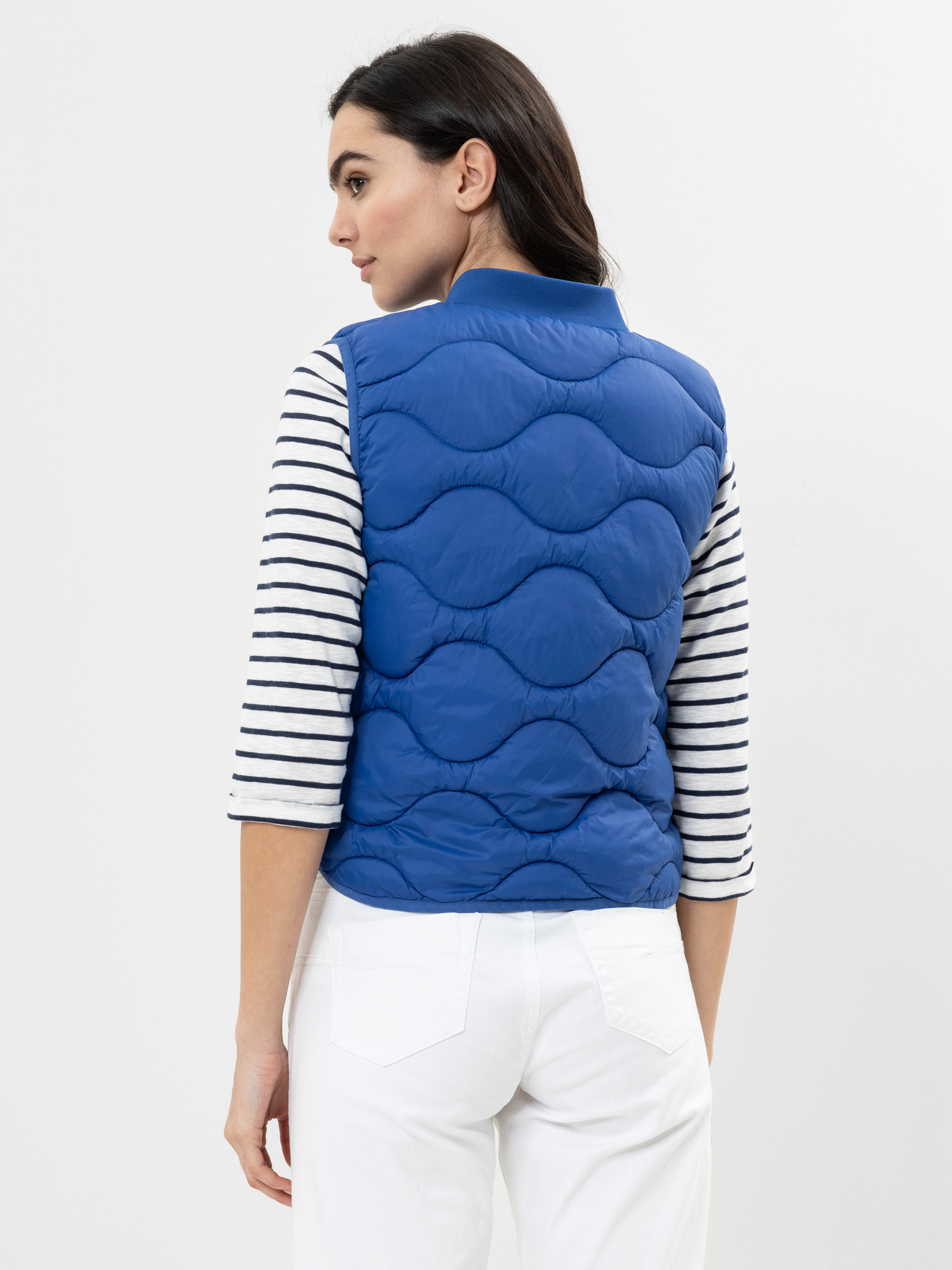 Eine Frau mit langen dunklen Haaren wird von hinten gezeigt. Sie trägt eine blaue Steppweste über einem weiß-schwarz gestreiften langärmeligen Hemd und einer weißen Hose und steht vor einem schlichten hellen Hintergrund.