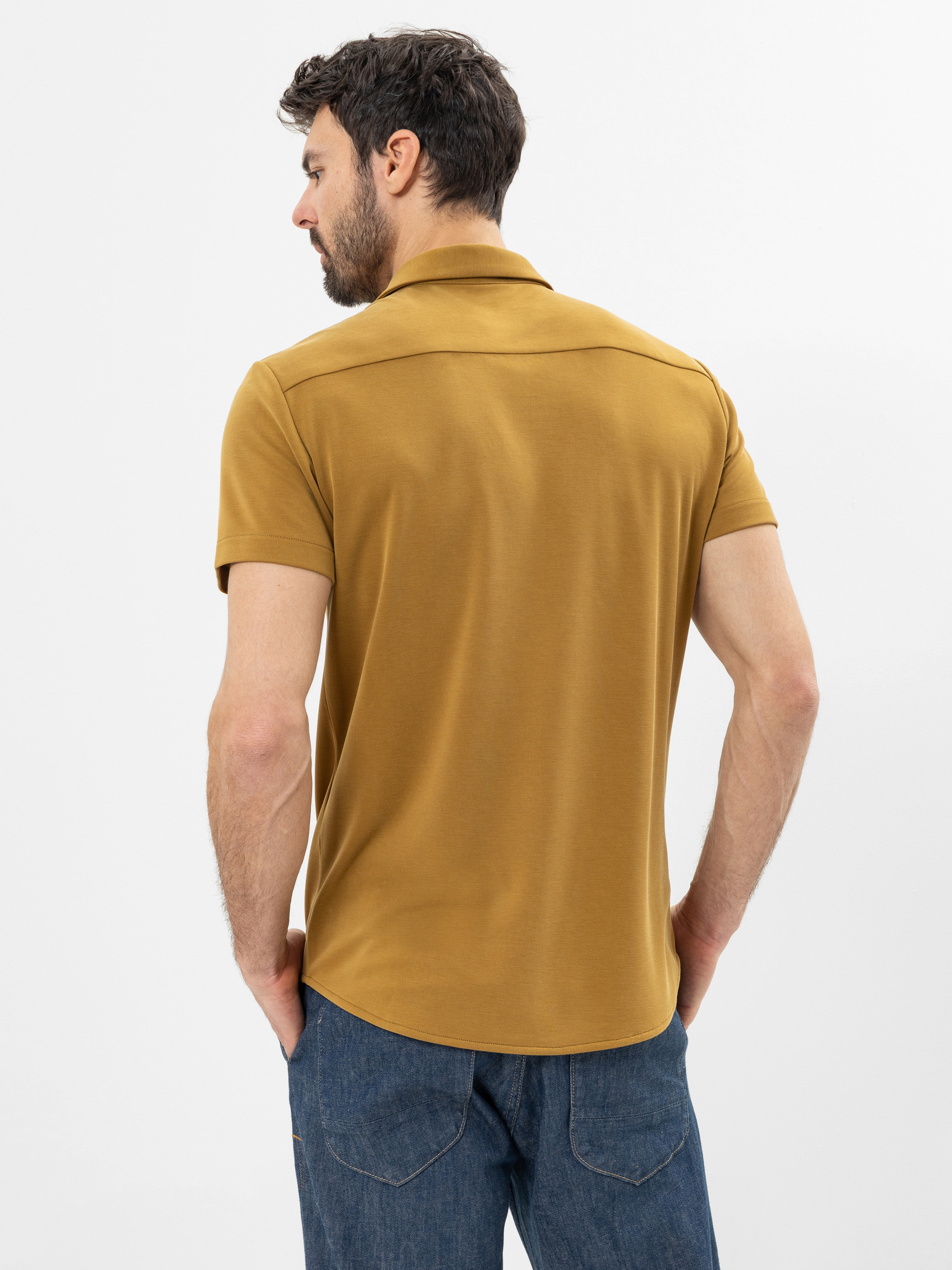 Ein Mann mit kurzen dunklen Haaren und Bart ist von hinten zu sehen. Er trägt ein senfgelbes, kurzärmeliges Poloshirt und blaue Jeans und steht vor einem schlichten weißen Hintergrund.