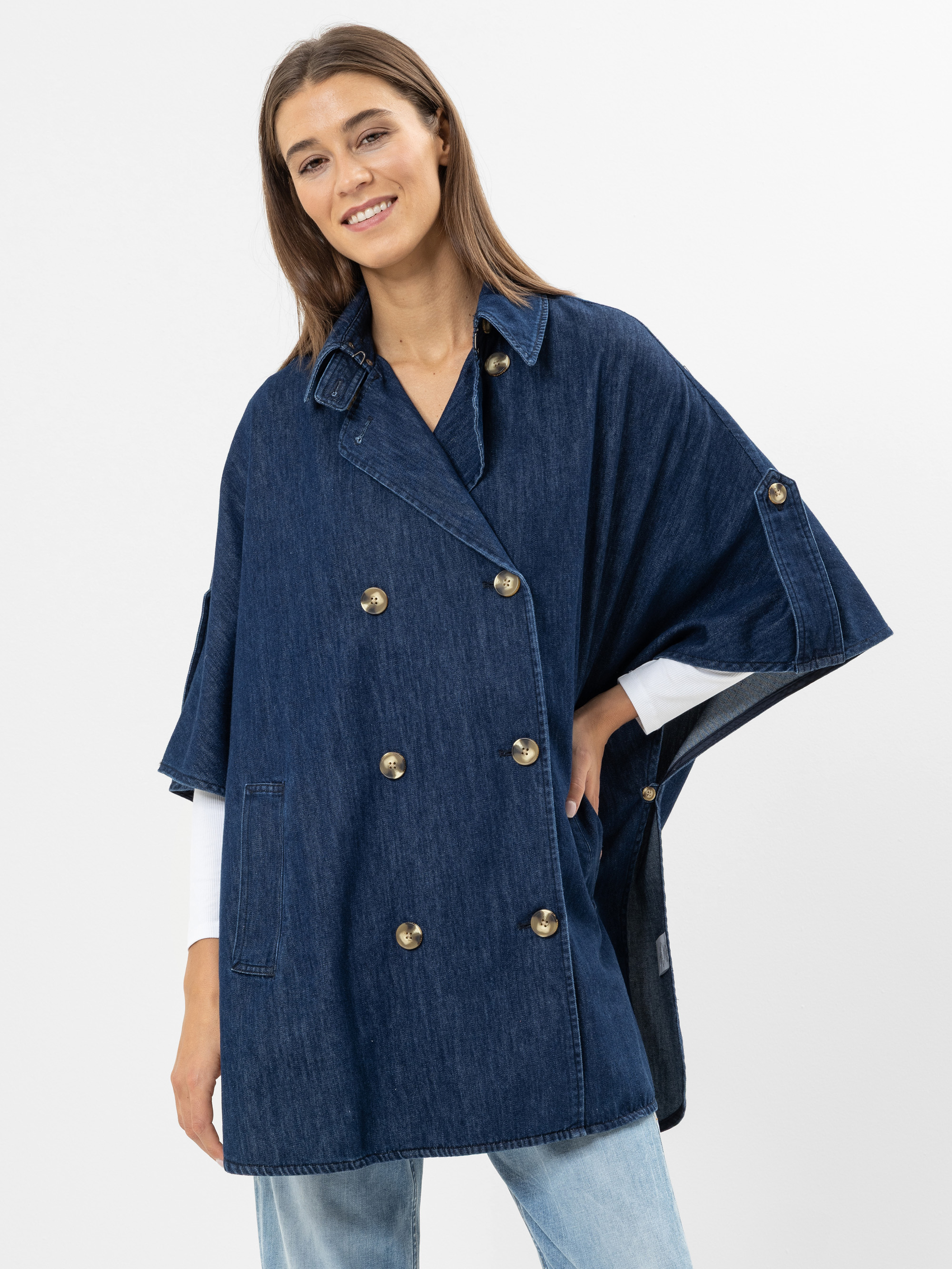 Eine Frau lächelt, während sie einen doppelreihigen Umhang aus dunklem Jeansstoff mit goldenen Knöpfen über einer weißen Bluse und hellblauen Jeans vor einem schlichten weißen Hintergrund trägt.