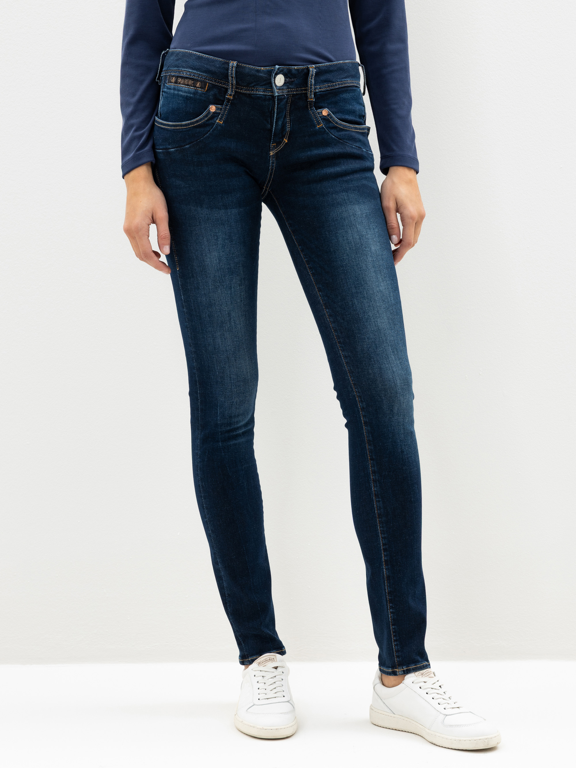 Eine Person, die dunkelblaue Röhrenjeans, ein langärmeliges marineblaues Oberteil und weiße Turnschuhe trägt, steht vor einem schlichten weißen Hintergrund. Das Bild zeigt die Person von den Schultern bis zu den Füßen.