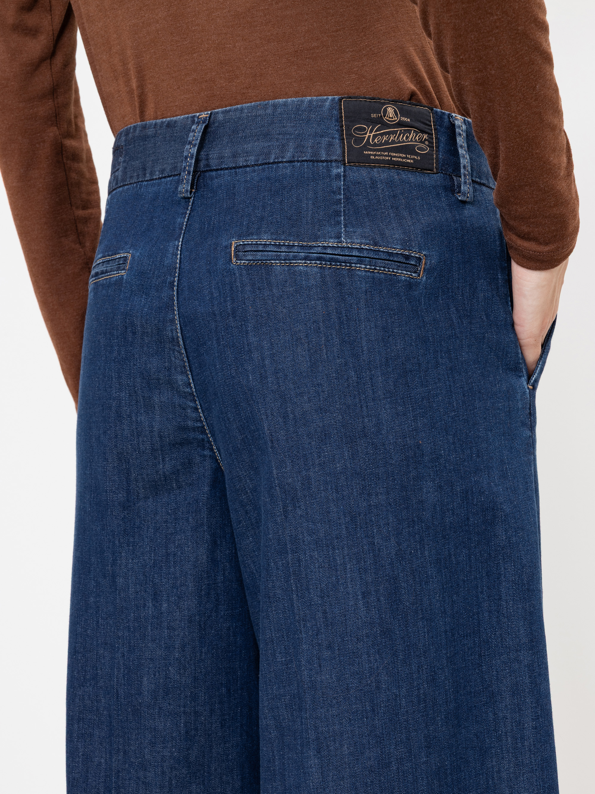 Eine Person, die eine blaue, weit geschnittene Jeanshose mit den Händen in den Taschen und ein braunes, langärmeliges Hemd mit einem schwarzen Etikett der Marke Henrietta am Bund trägt.