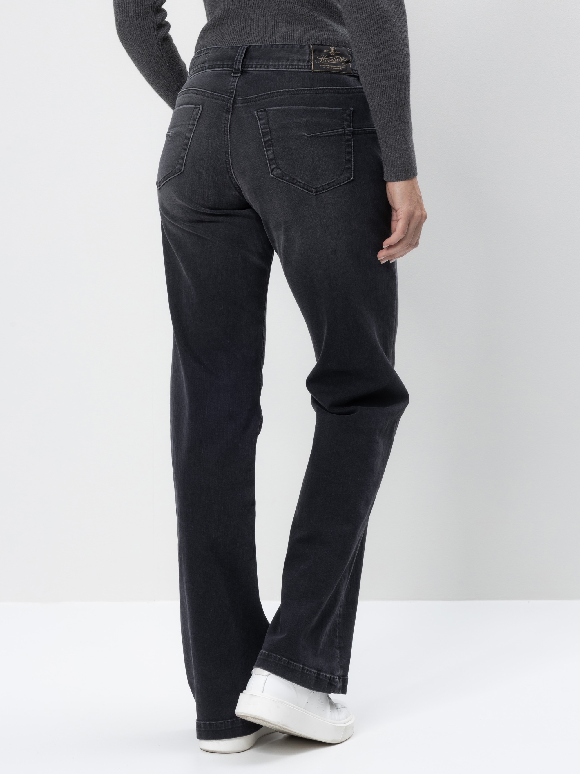 Eine Person, die dunkle Jeans mit geradem Bein, ein graues langärmeliges Oberteil und weiße Turnschuhe trägt, steht mit dem Gesicht zur Kamera vor einem schlichten weißen Hintergrund.
