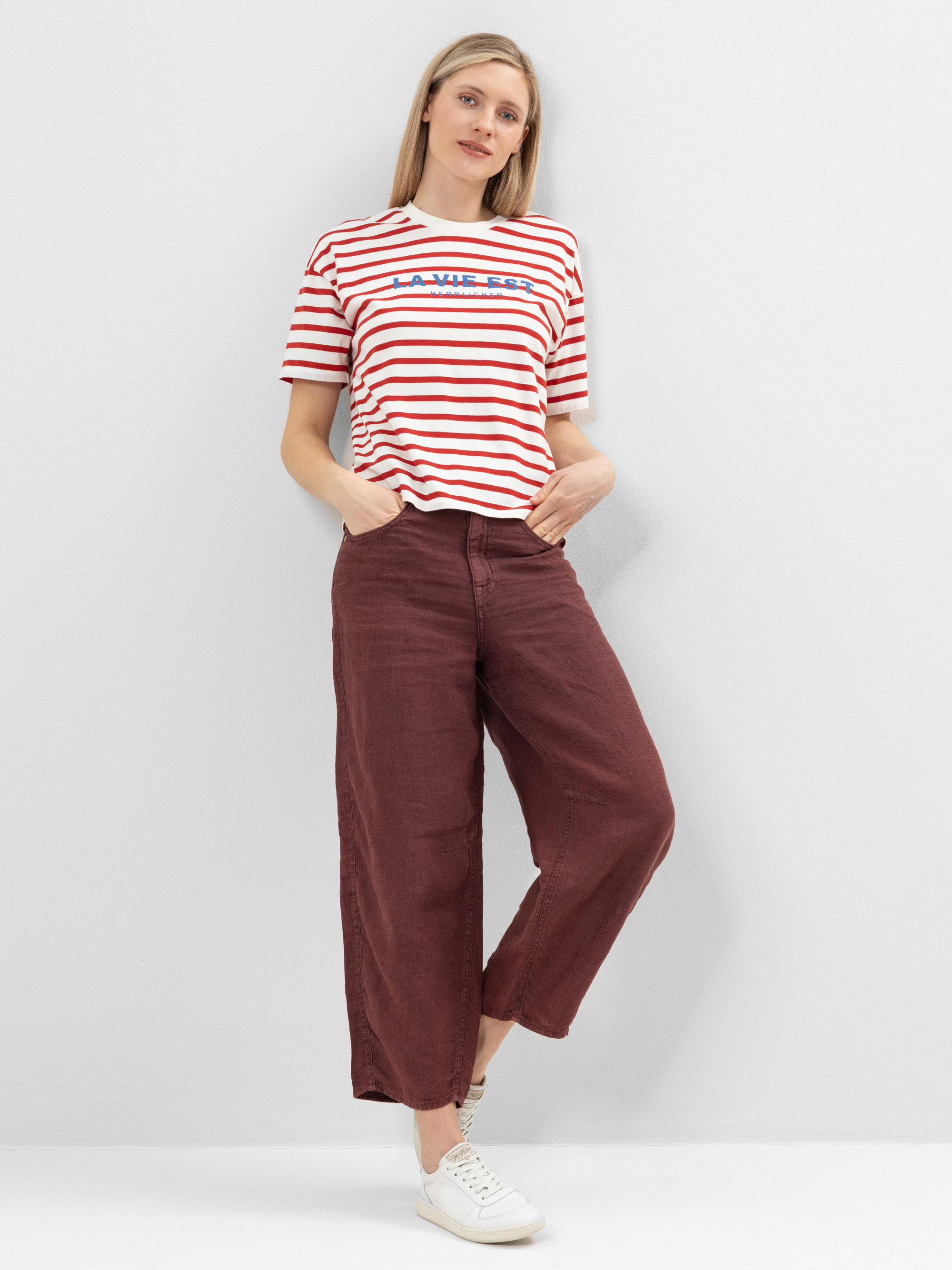 Eine Frau steht an einer weißen Wand. Sie trägt ein rot-weiß gestreiftes T-Shirt mit der Aufschrift LA VIE EST, eine kastanienbraune Hose mit weitem Bein und weiße Turnschuhe. Sie hat eine Hand in der Tasche und einen entspannten Gesichtsausdruck.