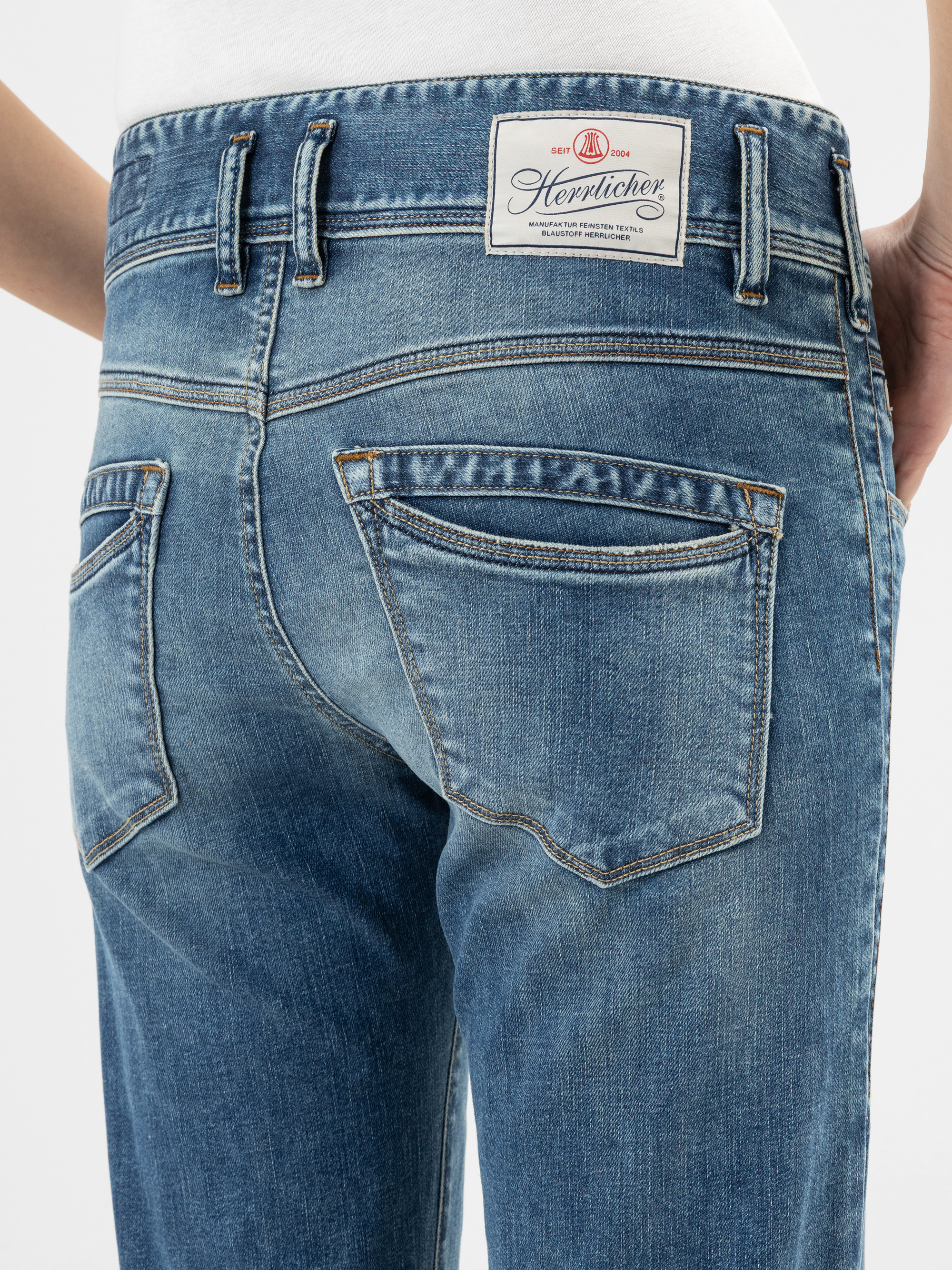 Nahaufnahme des Rückens einer Person, die eine blaue Denim-Jeans mit einem weißen Etikett am Bund trägt, auf dem Herrlicher steht. Die Jeans hat zwei Gesäßtaschen mit leichten Ausbleichungen und orangefarbenen Nähten.