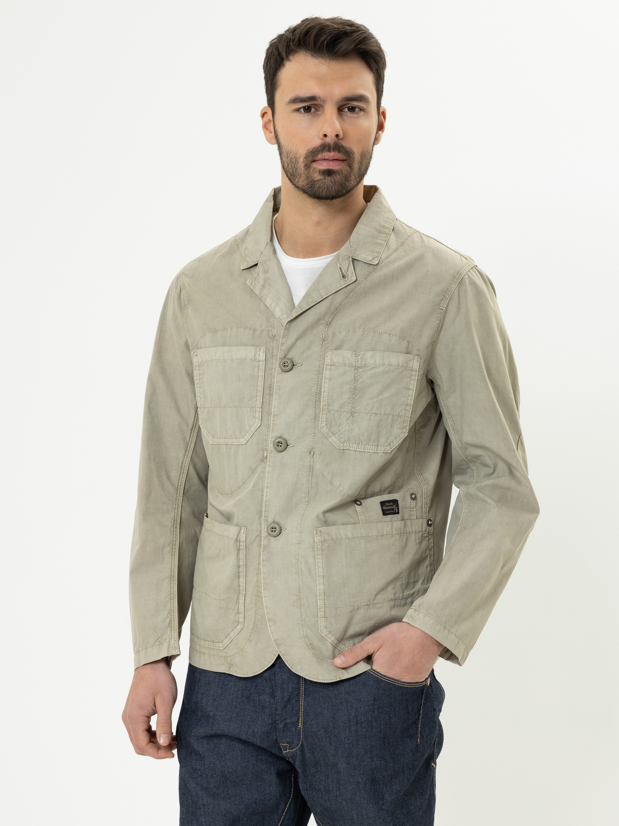 Ein Mann mit kurzen dunklen Haaren und einem Bart trägt eine hellbeige Freizeitjacke mit großen Vordertaschen und dunkle Jeans vor einem schlichten weißen Hintergrund.