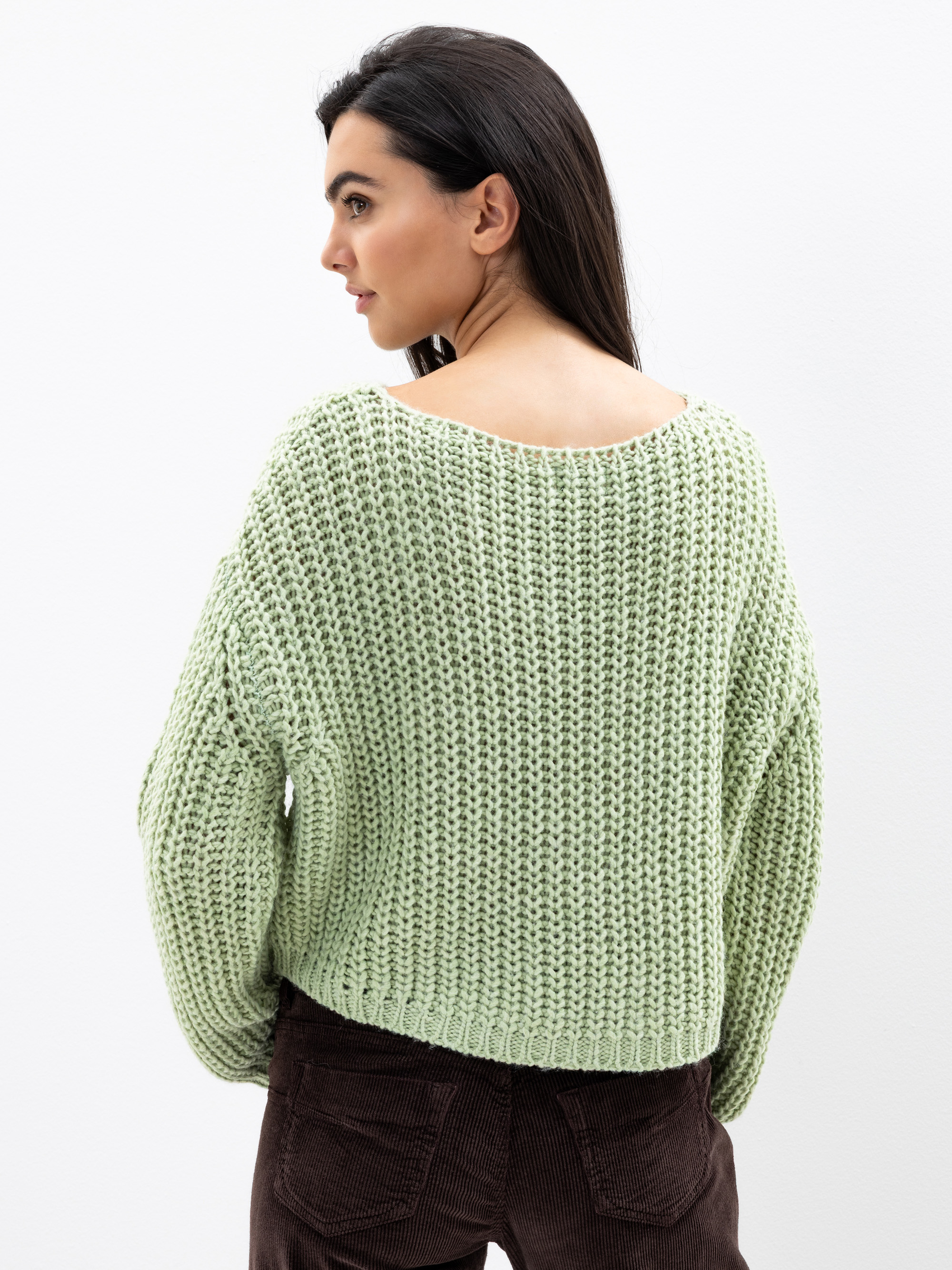 Eine Frau mit langen dunklen Haaren trägt einen hellgrünen Strickpulli und eine braune Hose. Sie blickt über die Schulter vor einem schlichten weißen Hintergrund.