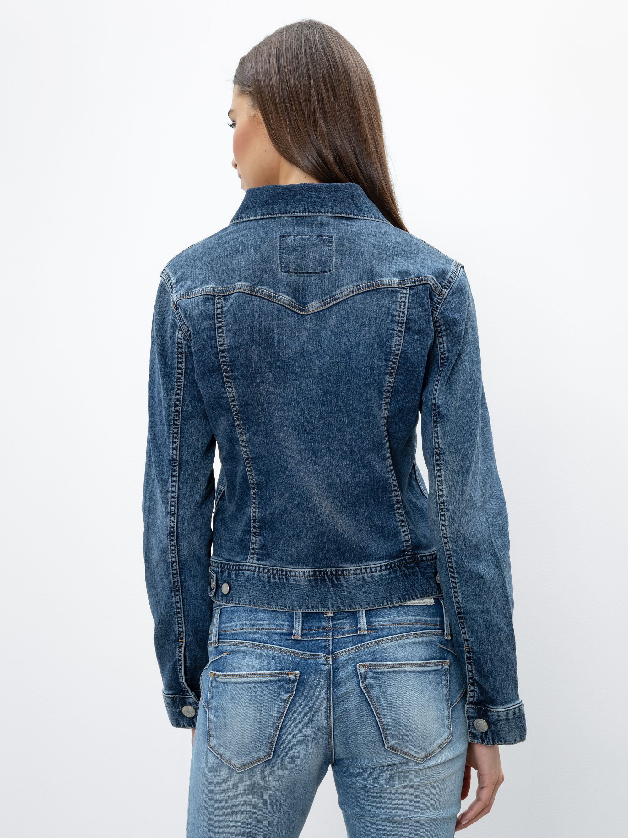 Eine Frau mit langen braunen Haaren, von hinten gesehen, trägt eine blaue Jeansjacke und hellblaue Jeans vor einem schlichten weißen Hintergrund.