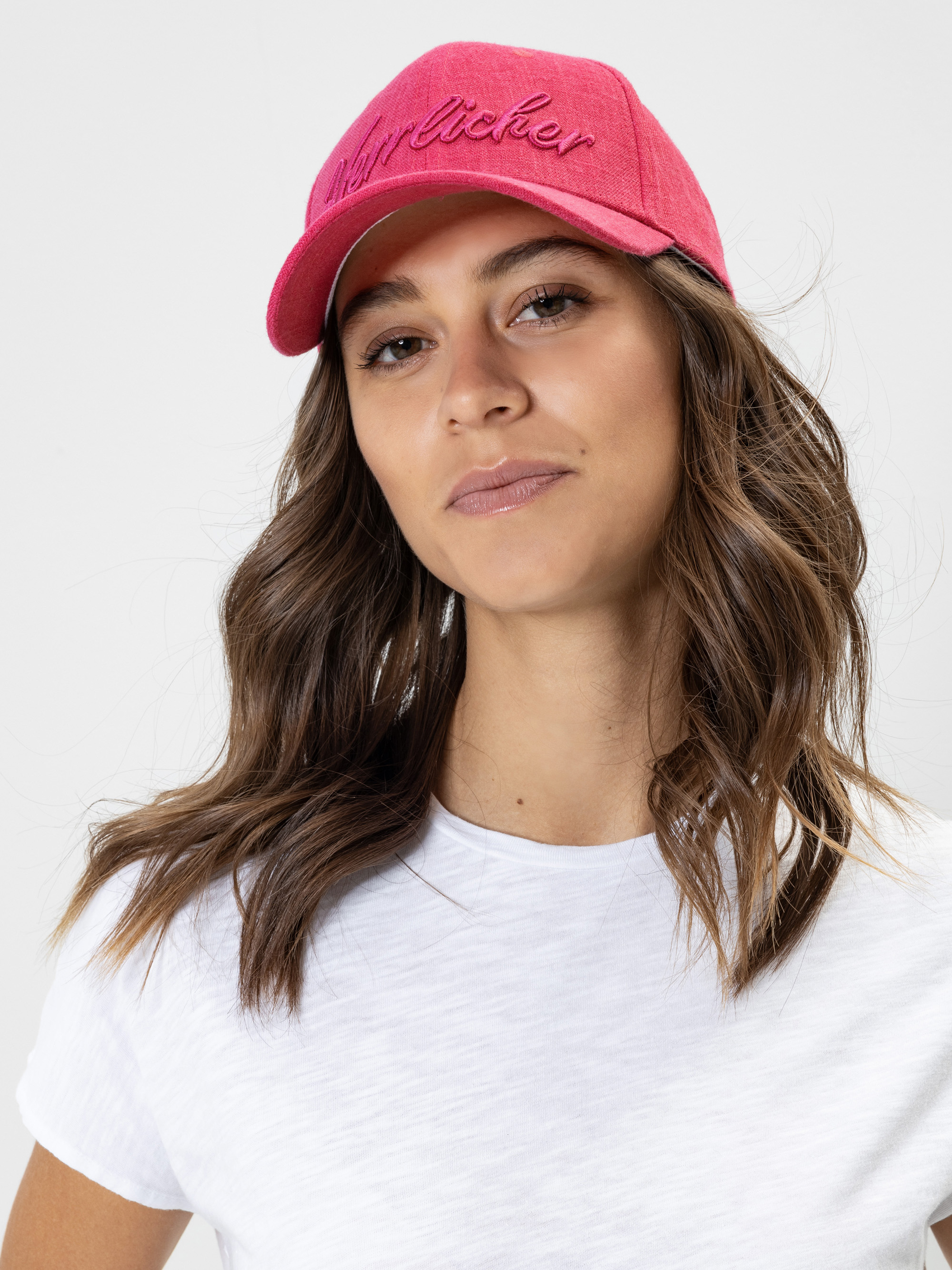 Eine Frau mit gewelltem braunem Haar trägt eine rosafarbene Baseballkappe und ein weißes T-Shirt und blickt selbstbewusst in die Kamera vor einem schlichten hellen Hintergrund.