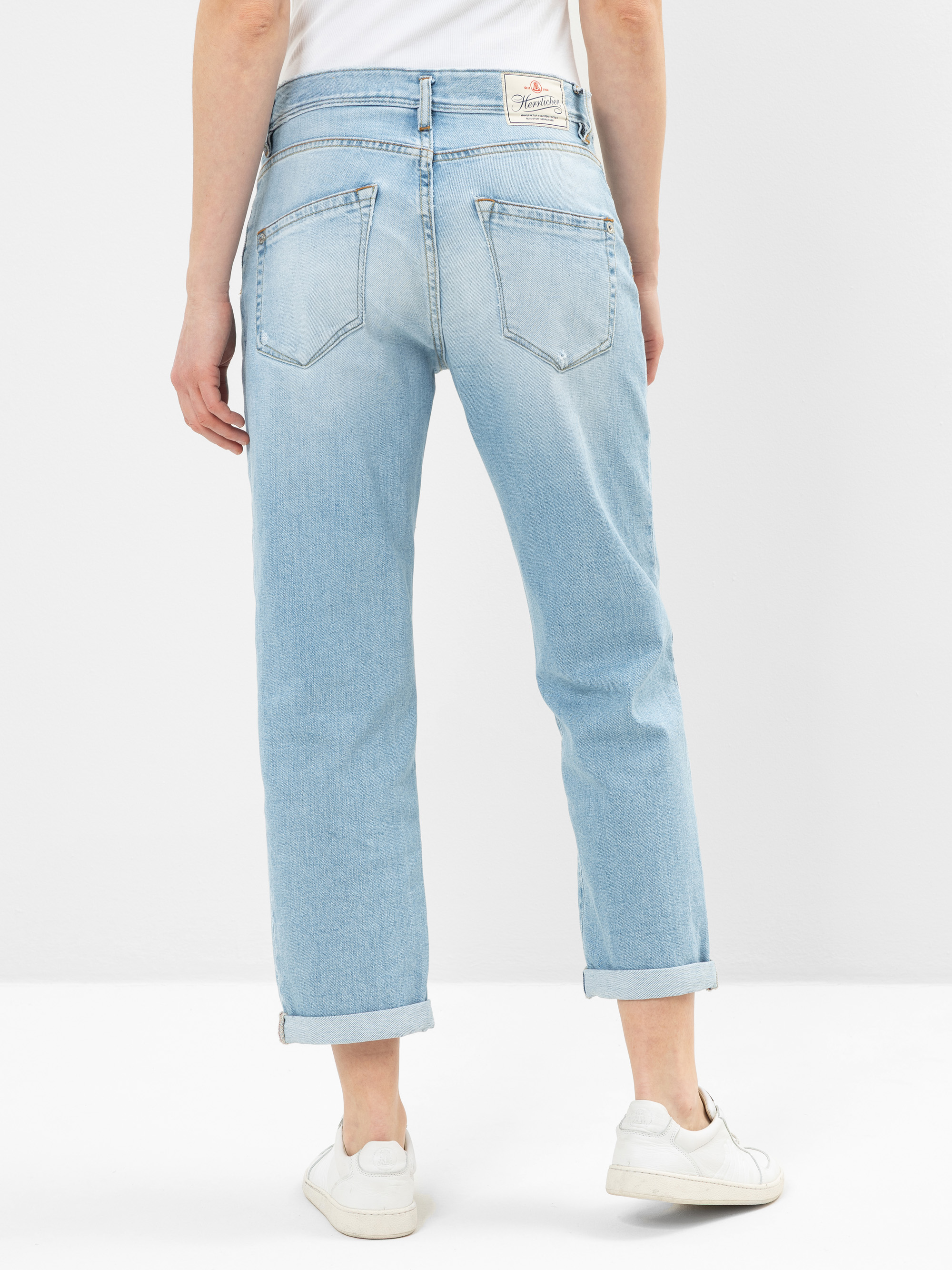 Eine Person, die hellblaue Jeans mit Bündchen und weiße Turnschuhe trägt, steht mit dem Rücken zur Kamera vor einem schlichten weißen Hintergrund. Die Jeans hat Gesäßtaschen und ein weißes Etikett am Bund.