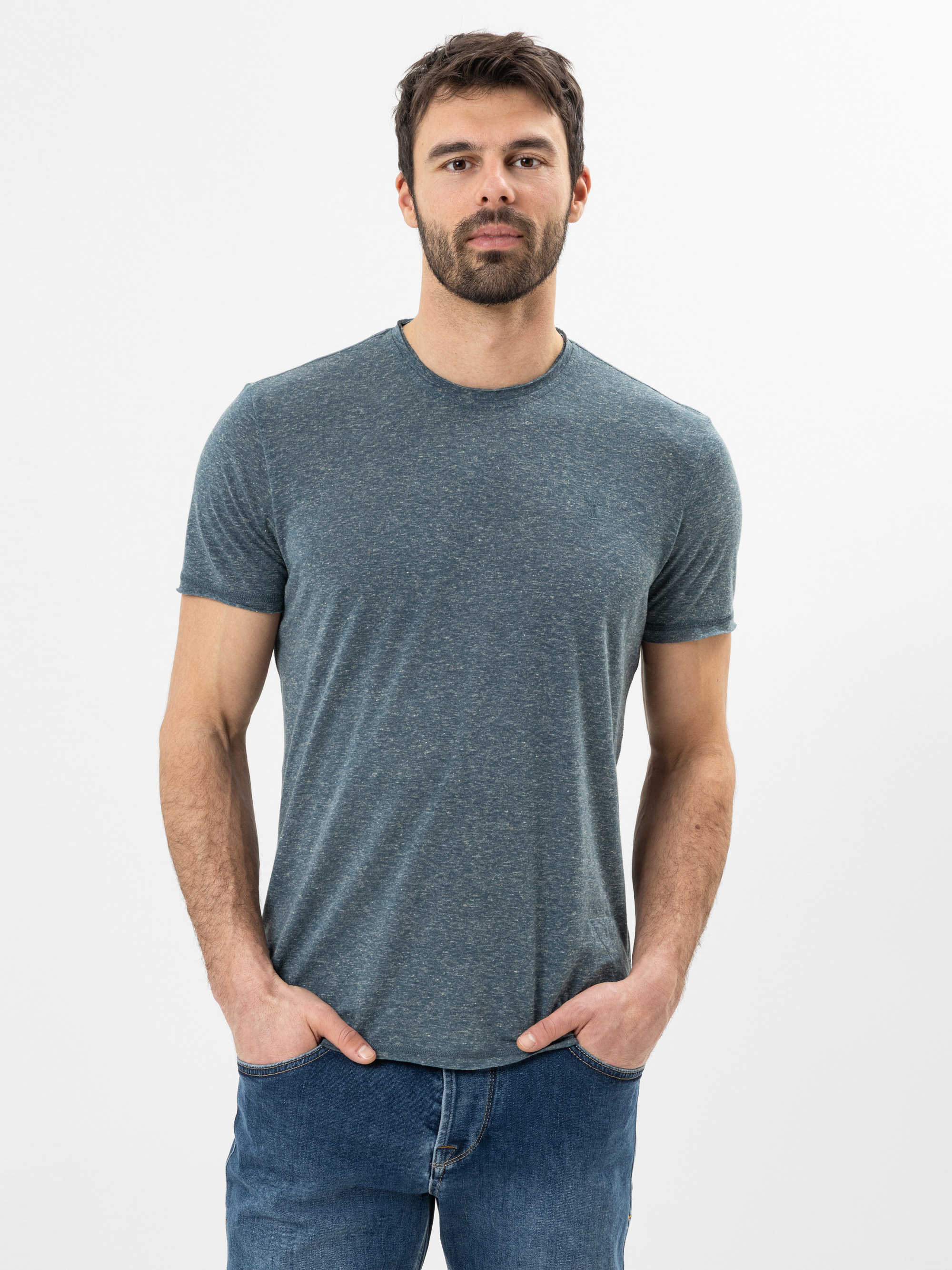 Ein Mann mit kurzen braunen Haaren und einem Bart trägt ein tailliertes, kurzärmeliges, blaugraues T-Shirt und blaue Jeans. Er steht vor einem schlichten weißen Hintergrund und hat die Hände in den Taschen.