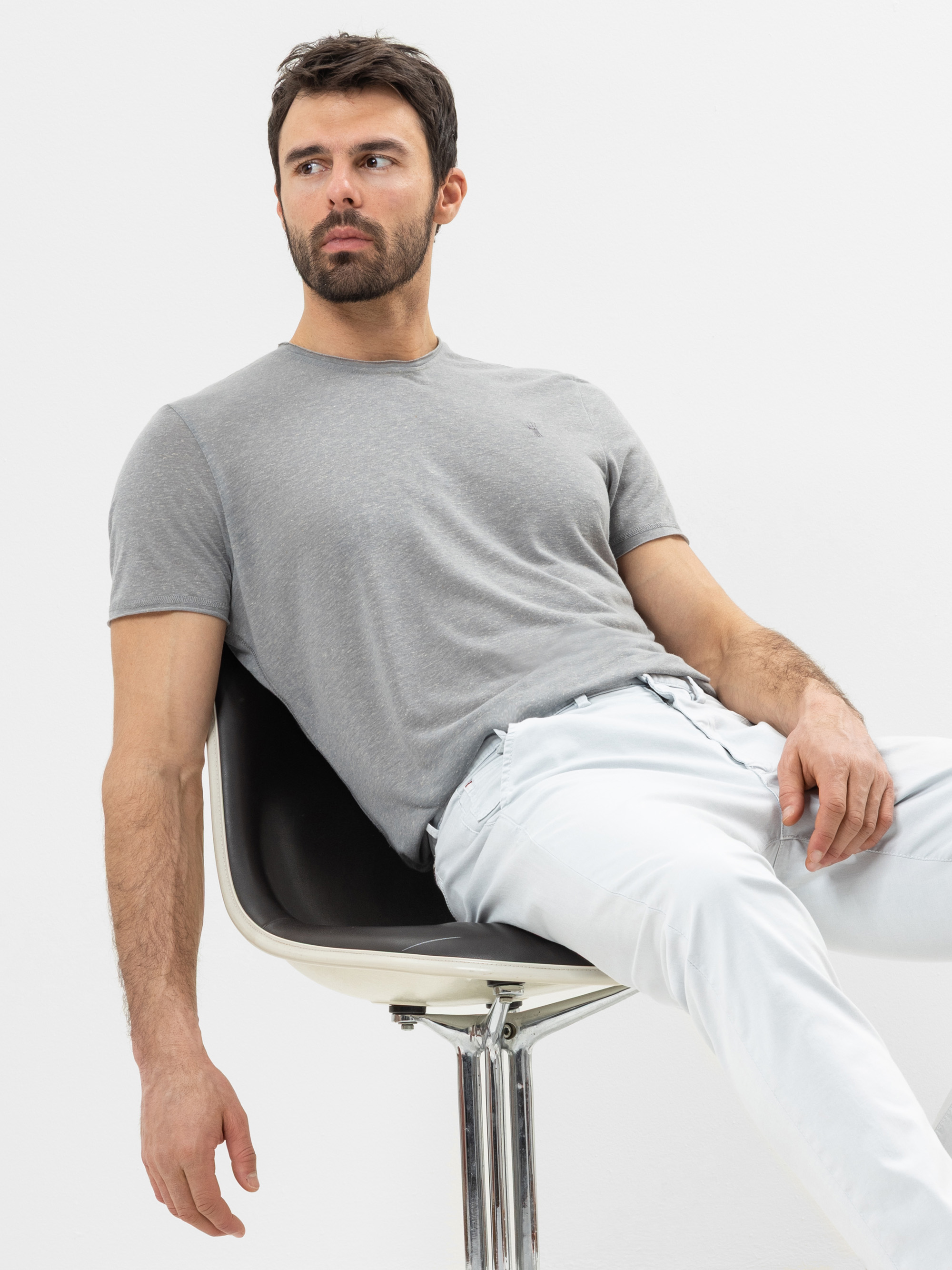 Ein Mann mit dunklem Haar und Bart sitzt lässig auf einem schwarz-weißen Stuhl, trägt ein hellgraues T-Shirt und eine weiße Hose, vor einem schlichten weißen Hintergrund.