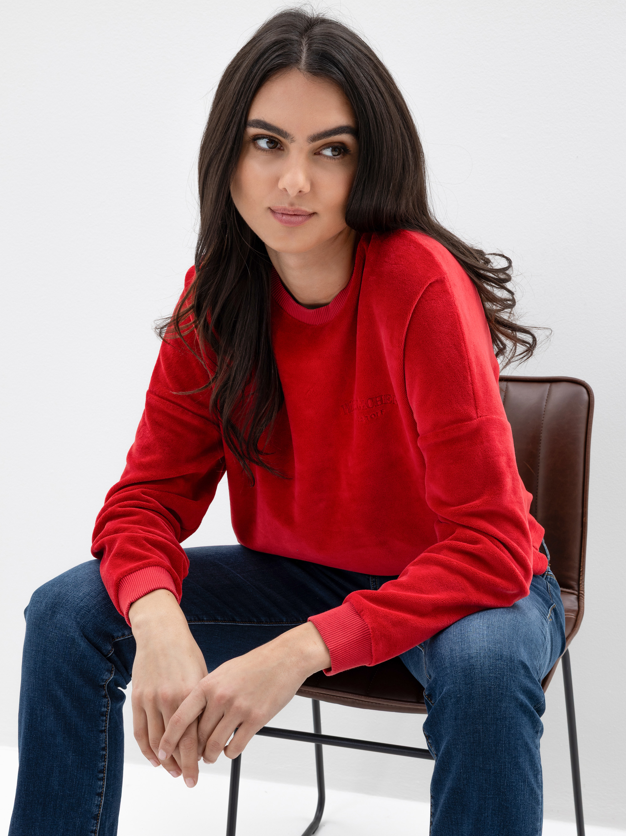 Eine Frau mit langen dunklen Haaren, die ein rotes Sweatshirt und blaue Jeans trägt, sitzt auf einem braunen Stuhl vor einem schlichten weißen Hintergrund und blickt mit neutralem Blick leicht zur Seite.