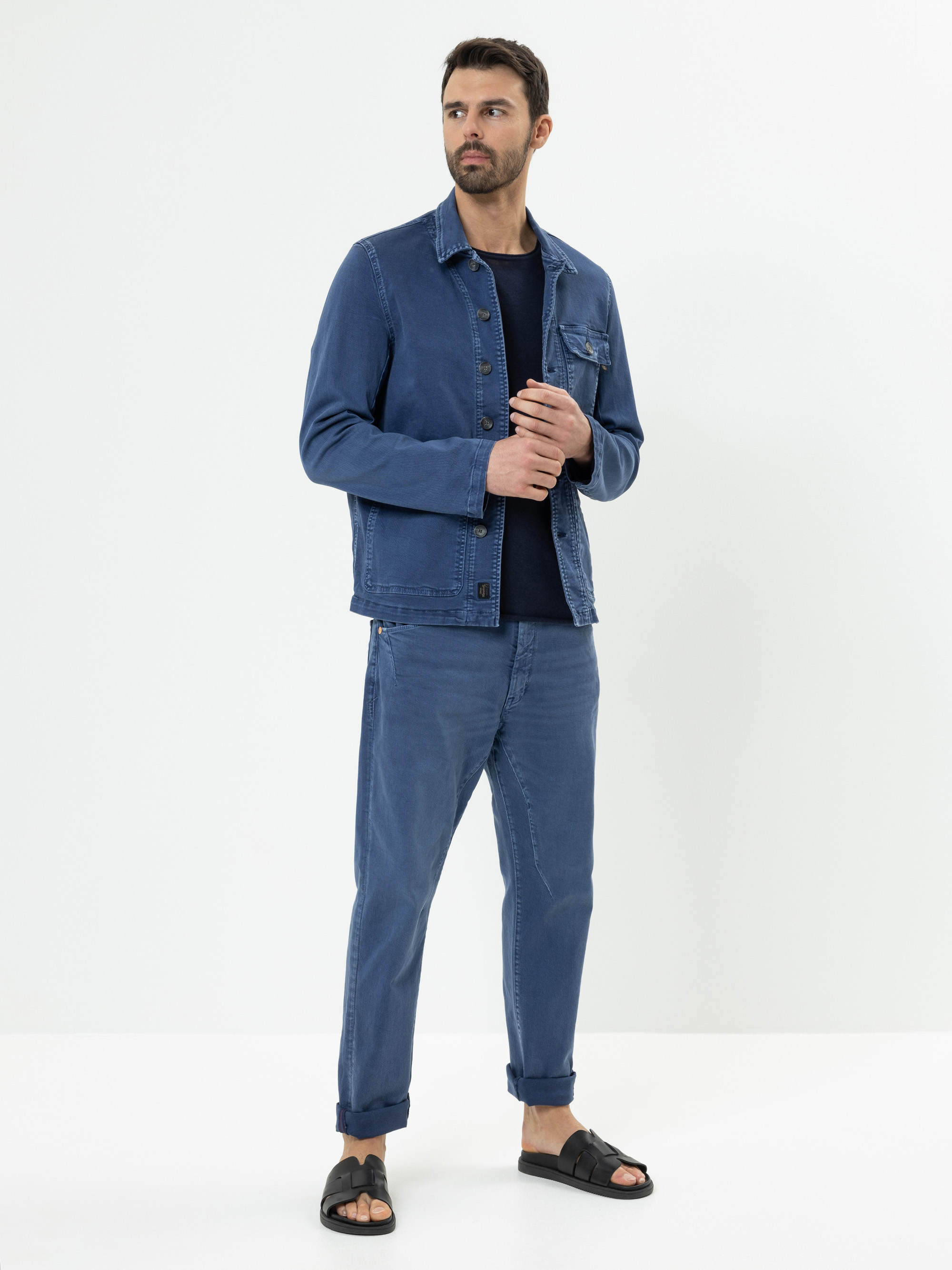 Ein Mann steht vor einem schlichten weißen Hintergrund und trägt eine blaue Jeansjacke, dazu passende blaue Jeans, ein schwarzes Hemd und schwarze Sandalen. Er schaut leicht zur Seite und hat einen neutralen Blick.