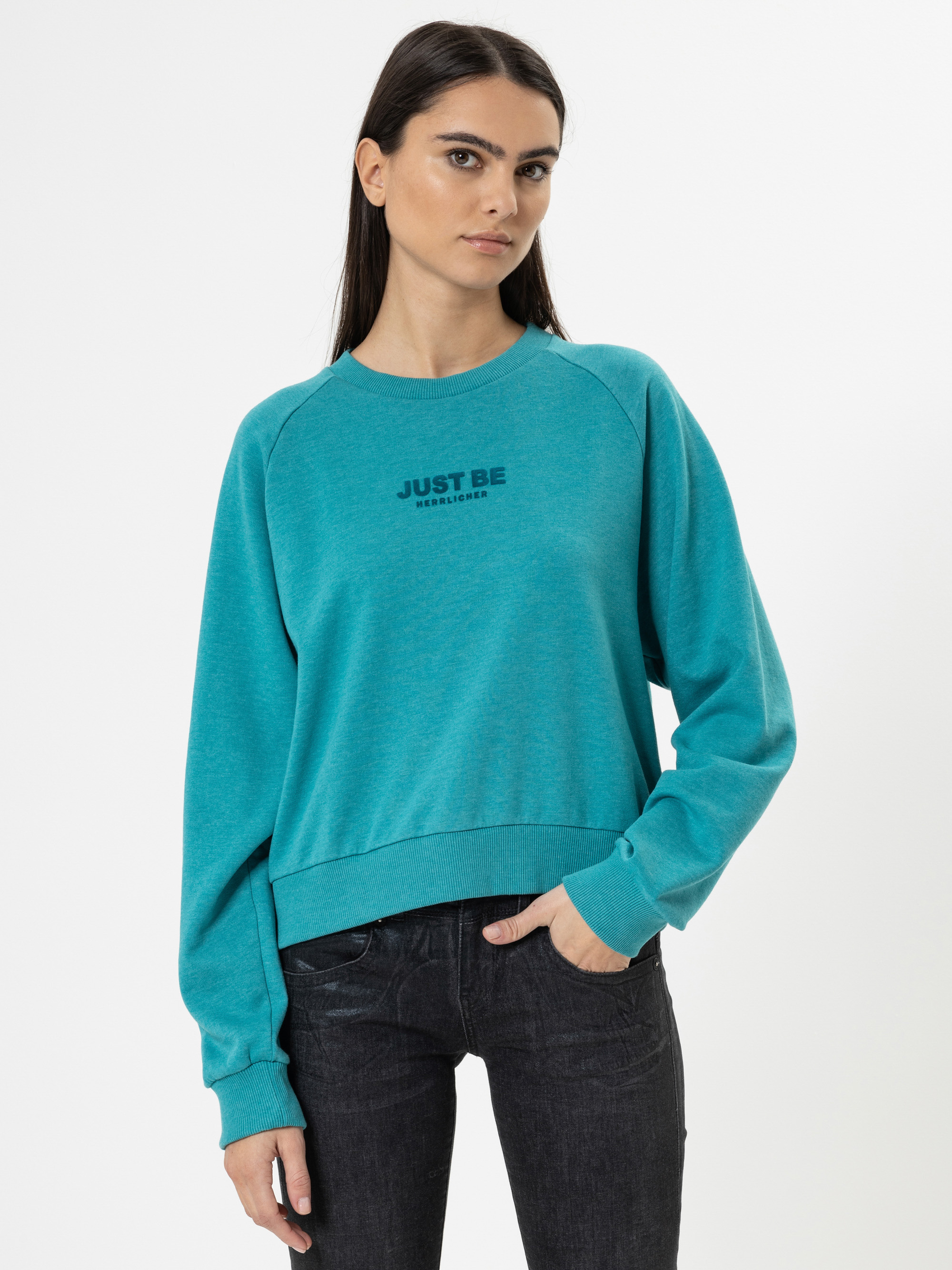 Eine Frau mit langen dunklen Haaren trägt ein türkisfarbenes Sweatshirt mit dem Aufdruck JUST BE REALWEAR auf der Vorderseite, gepaart mit einer dunklen Jeans, vor einem schlichten weißen Hintergrund.