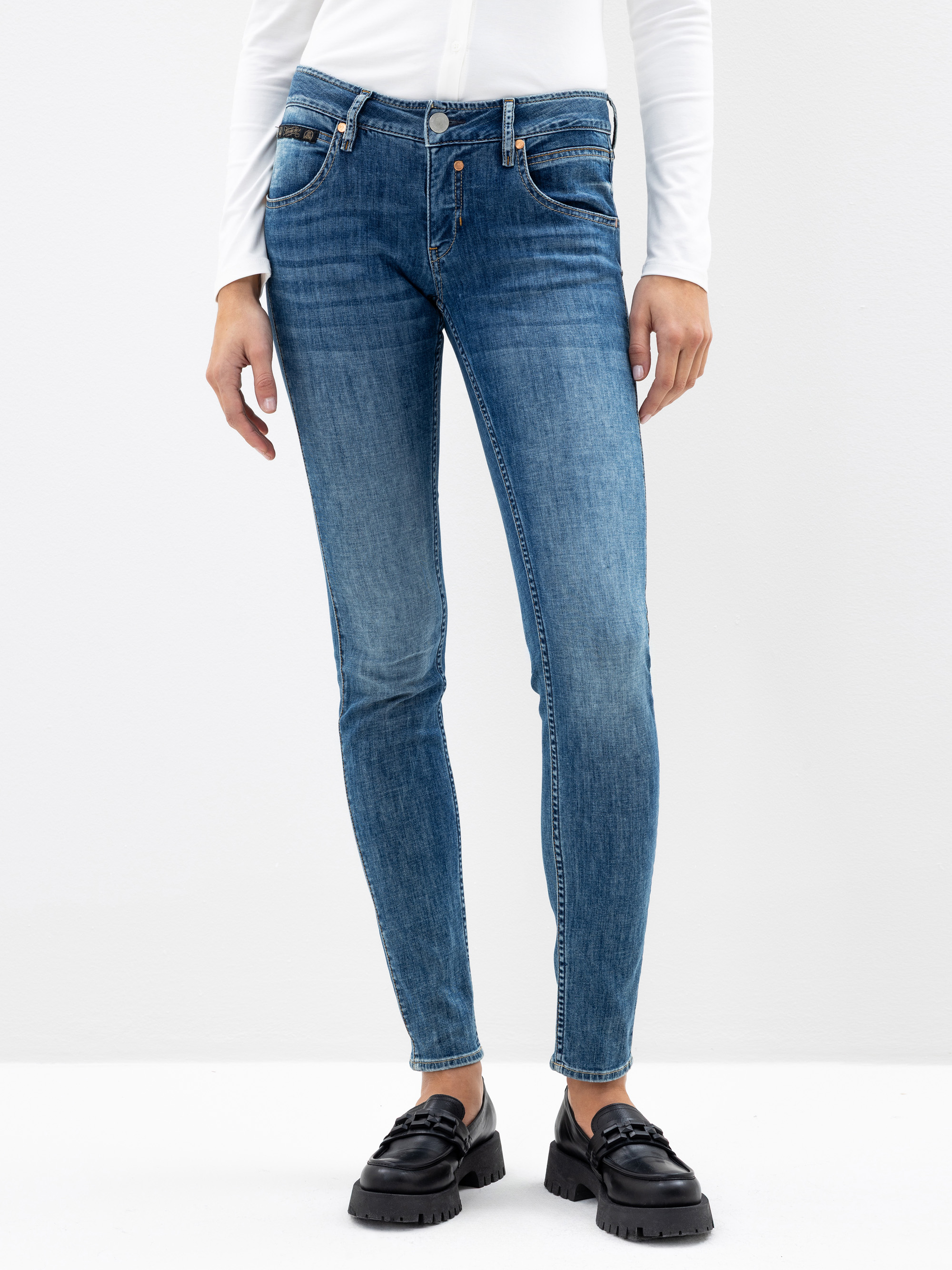 Eine Person in blauen Skinny-Jeans, einem weißen langärmeligen Hemd und schwarzen Halbschuhen steht vor einem schlichten weißen Hintergrund. Das Bild zeigt die Person von den Schultern bis zu den Füßen.