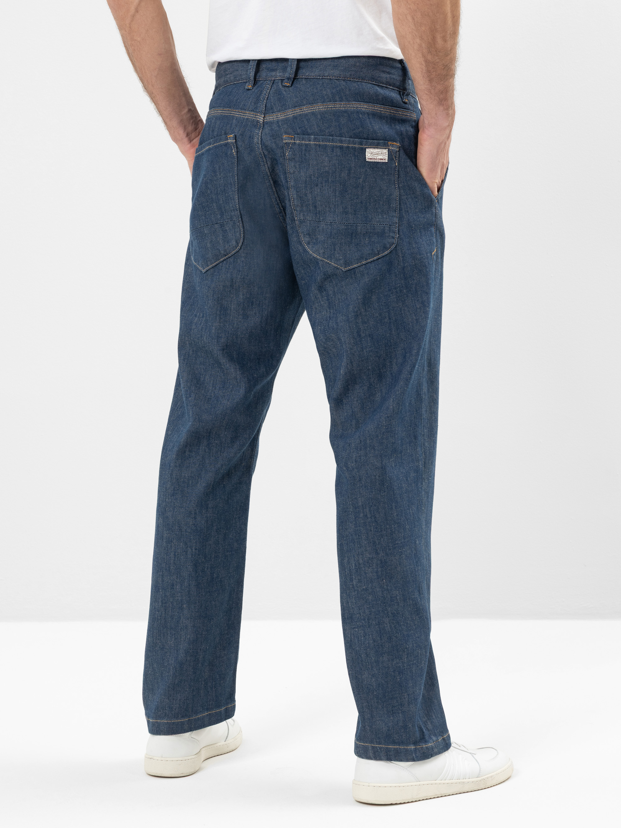 Mann in blauen Jeans mit geradem Bein und weißen Turnschuhen, stehend mit den Händen in den Hosentaschen, von der Kamera abgewandt, vor einem schlichten weißen Hintergrund.