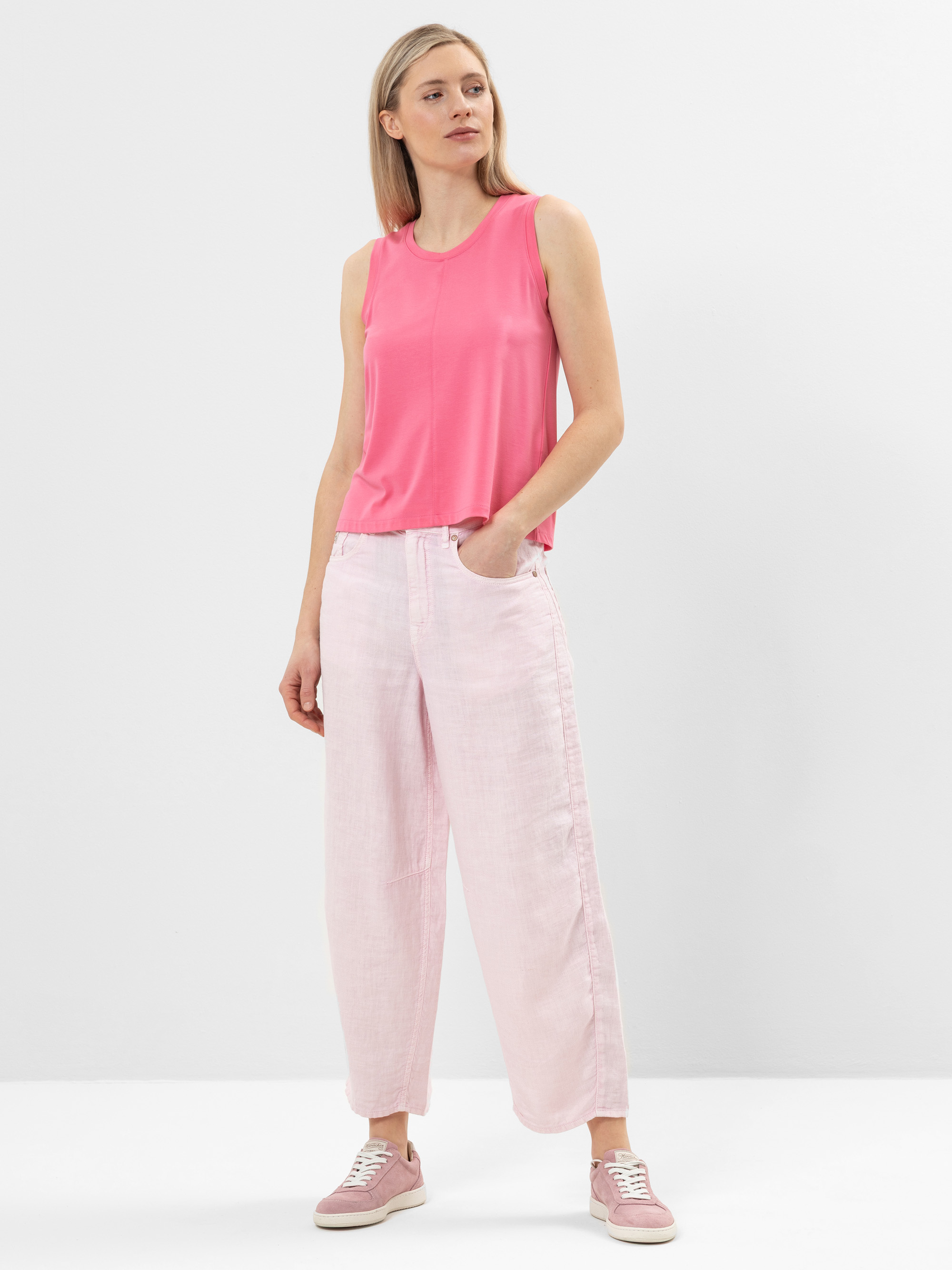 Eine Frau steht vor einem schlichten weißen Hintergrund und trägt ein hellrosa ärmelloses Oberteil, eine hellrosa Hose mit weitem Bein und rosa Turnschuhe. Sie hat helles Haar und eine Hand in der Tasche, wobei sie leicht zur Seite schaut.