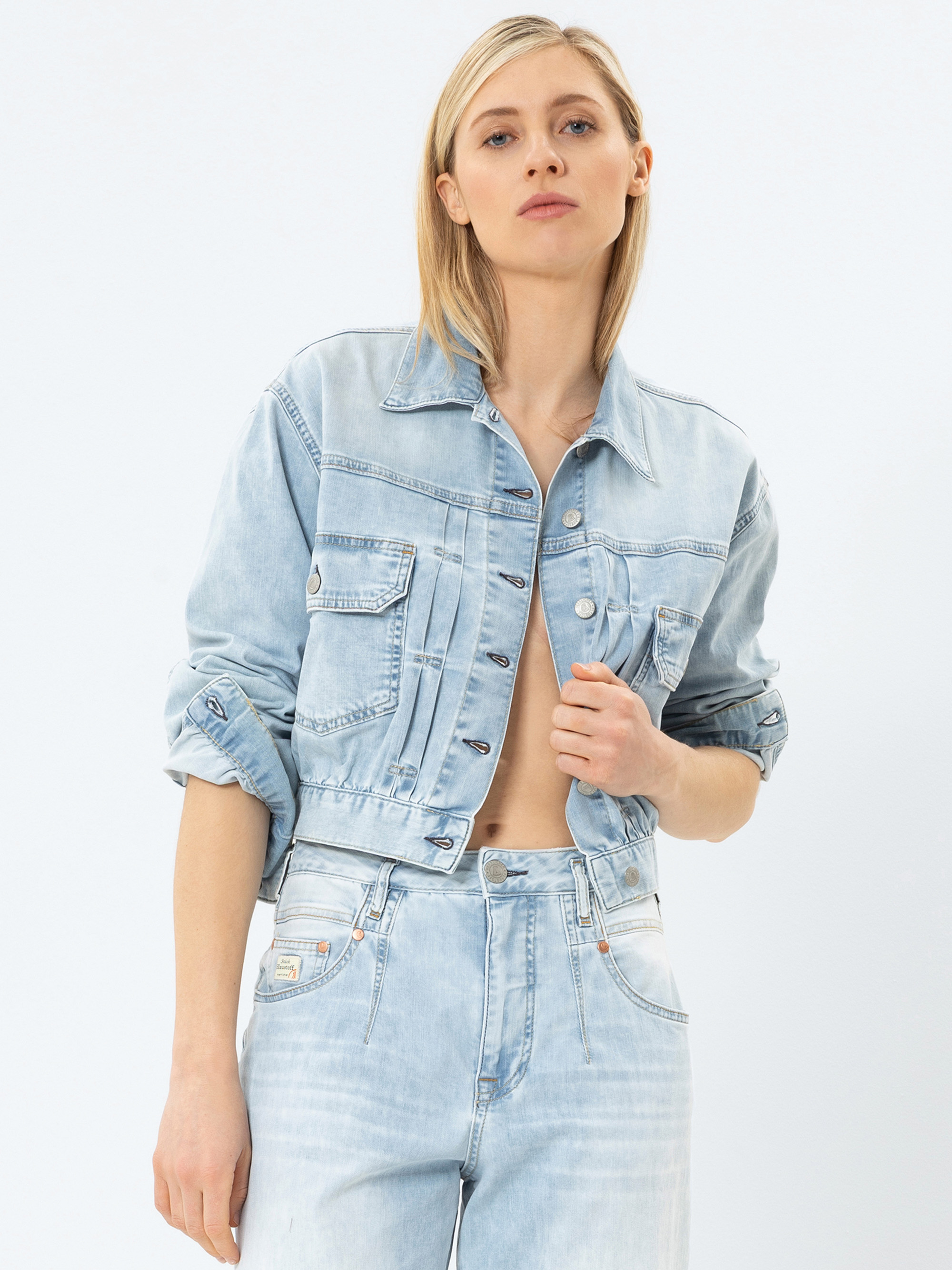 Eine Person mit glattem blondem Haar trägt eine hellblaue Jeansjacke und eine Jeans mit hoher Taille. Die Jacke ist oben zugeknöpft und die Ärmel sind hochgekrempelt. Der Hintergrund ist einfarbig weiß.