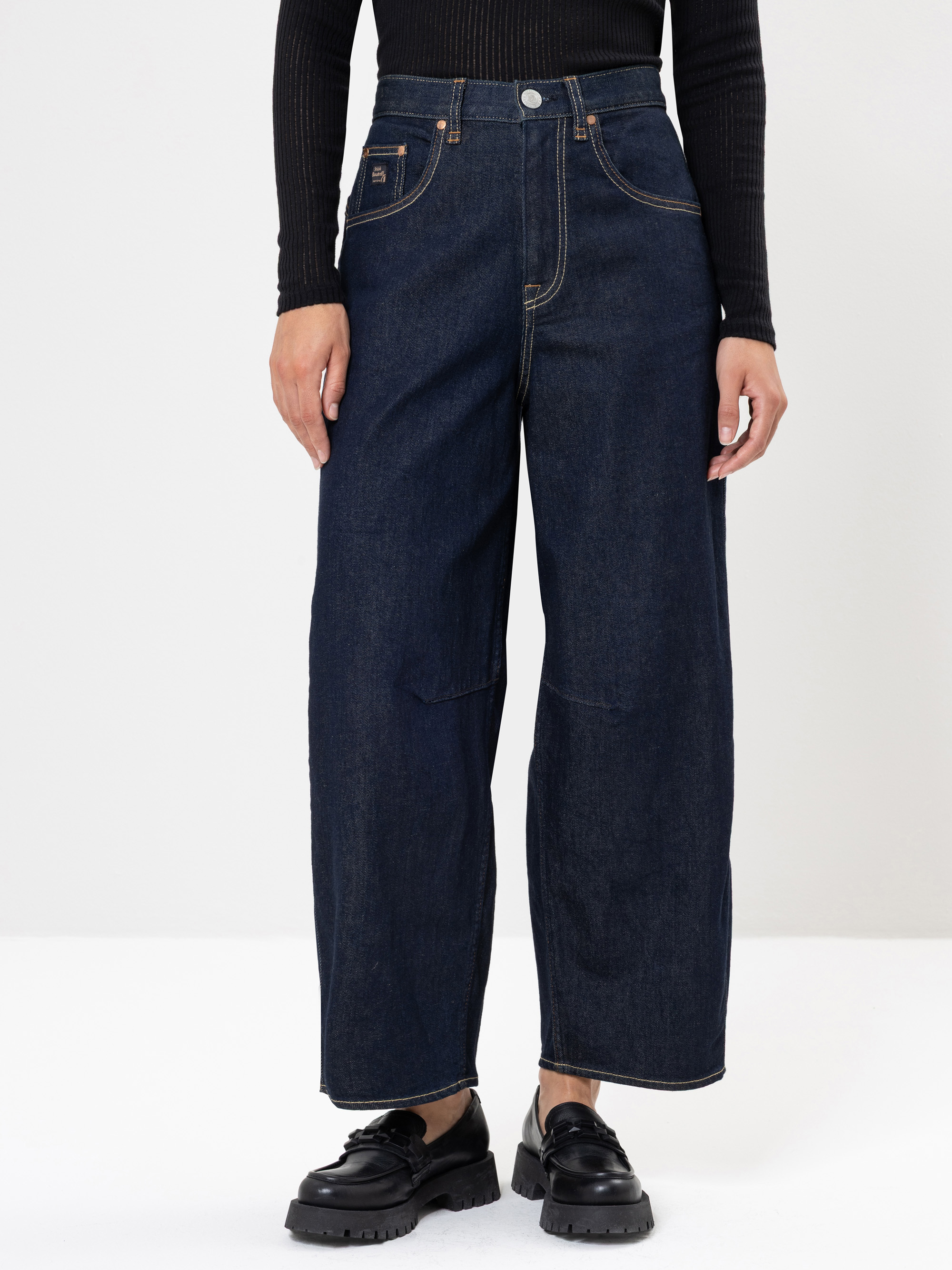 Person, die ein schwarzes, langärmeliges Oberteil, hochgeschnittene, weit geschnittene dunkelblaue Jeans und schwarze, klobige Schuhe trägt und vor einem schlichten, hellen Hintergrund steht. Das Gesicht ist auf dem Bild nicht zu sehen.