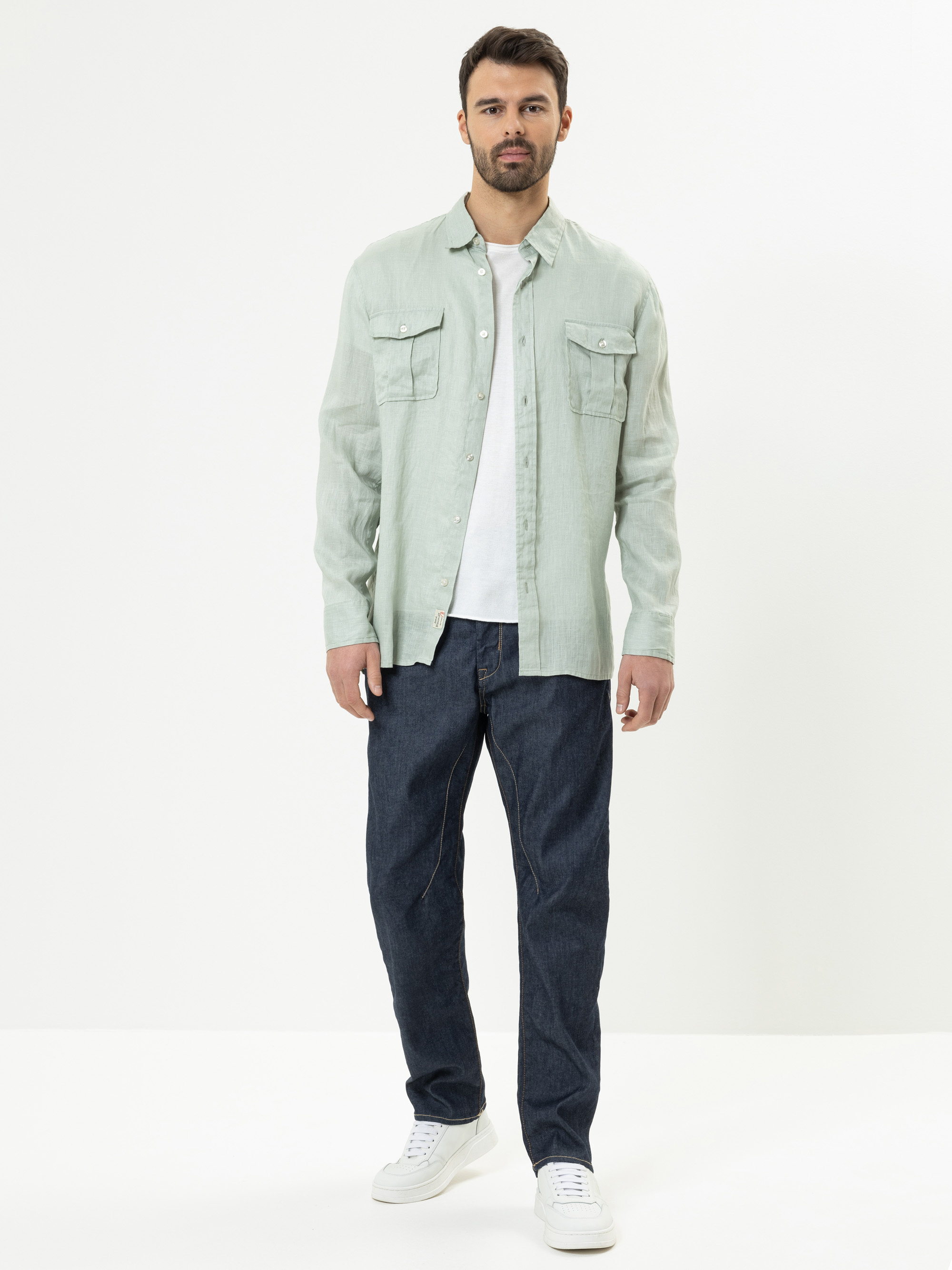Ein Mann steht vor einem schlichten weißen Hintergrund und trägt ein hellgrünes Button-up-Hemd über einem weißen T-Shirt, dunkelblaue Jeans und weiße Turnschuhe.