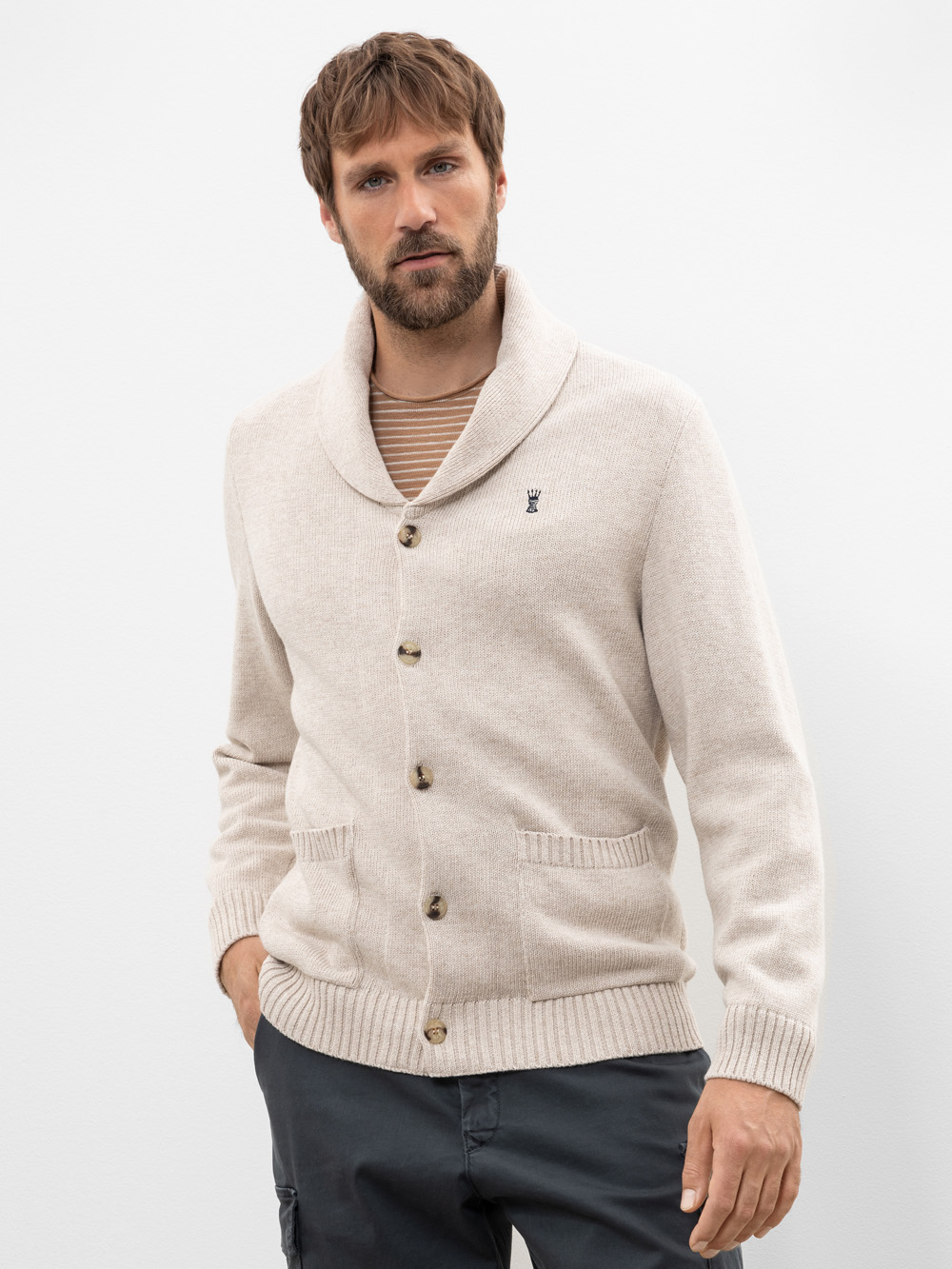 Ein Mann mit hellbraunem Haar und Bart trägt eine beigefarbene Strickjacke mit Schalkragen und braunen Knöpfen über einem gestreiften Hemd. Er steht vor einem schlichten weißen Hintergrund und hat eine Hand in der Tasche.