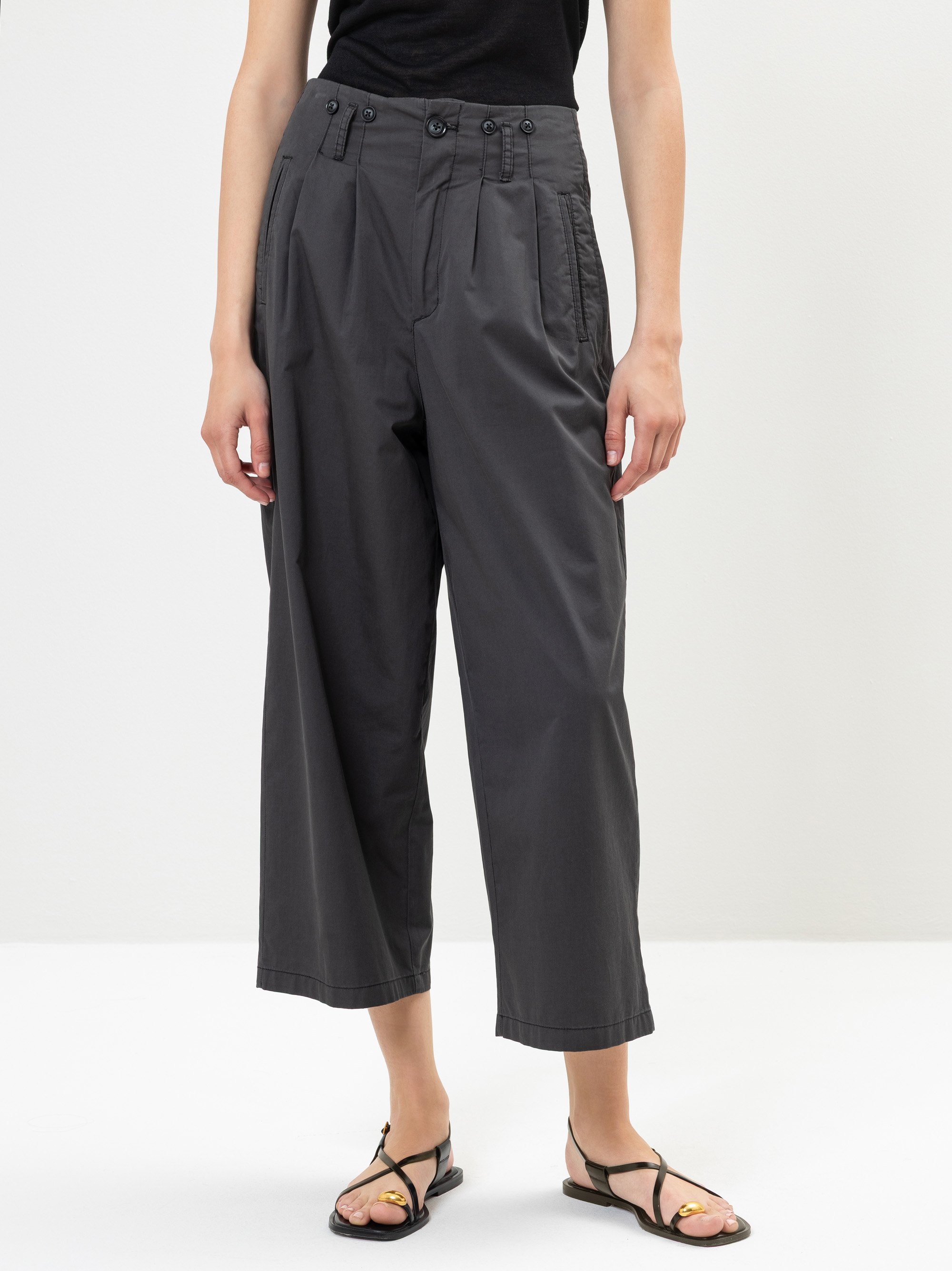 Eine Person, die ein schwarzes Oberteil, eine hochtaillierte dunkelgraue Hose mit weitem Bein, Falten und Knöpfen sowie schwarze Sandalen trägt, steht vor einem schlichten weißen Hintergrund.