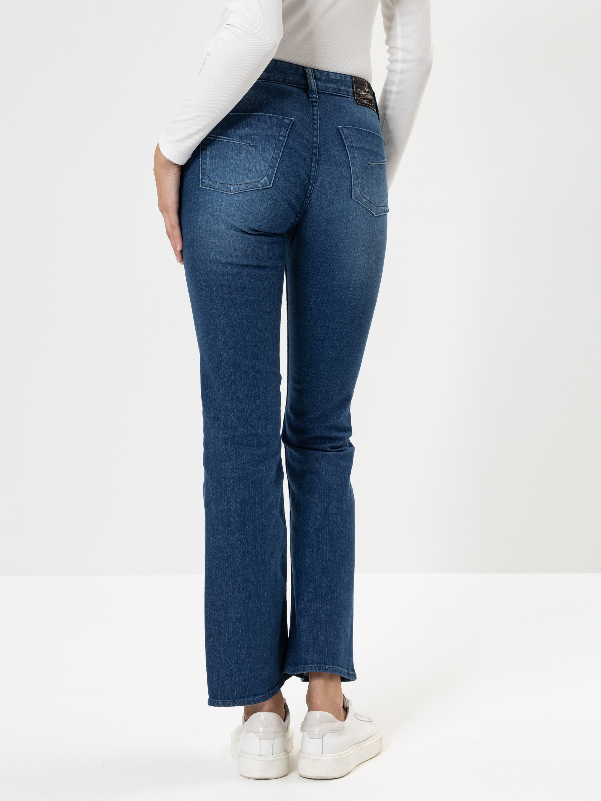 Eine Person, die blaue Jeans, ein weißes langärmeliges Oberteil und weiße Turnschuhe trägt, steht mit dem Rücken zur Kamera vor einem schlichten weißen Hintergrund.