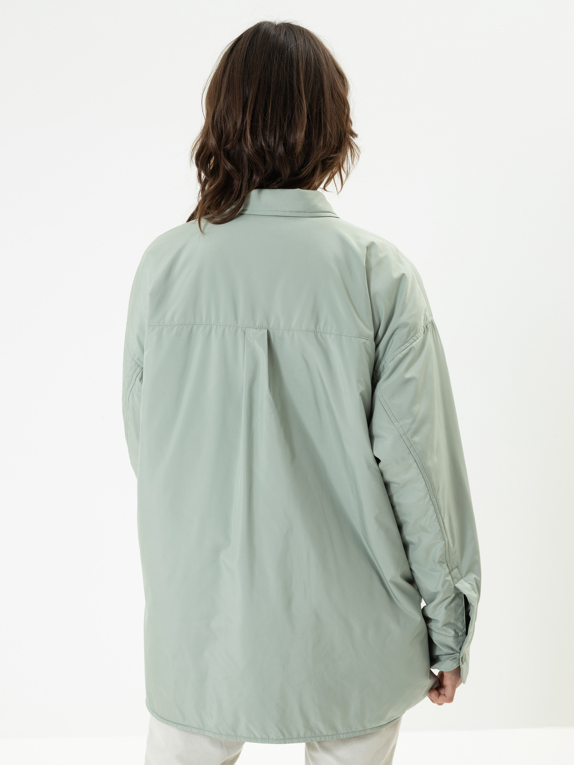 Eine Person mit schulterlangen braunen Haaren steht mit dem Rücken zur Kamera. Sie trägt eine lockere, hellgrüne Button-up-Jacke und eine cremefarbene Hose vor einem schlichten weißen Hintergrund.