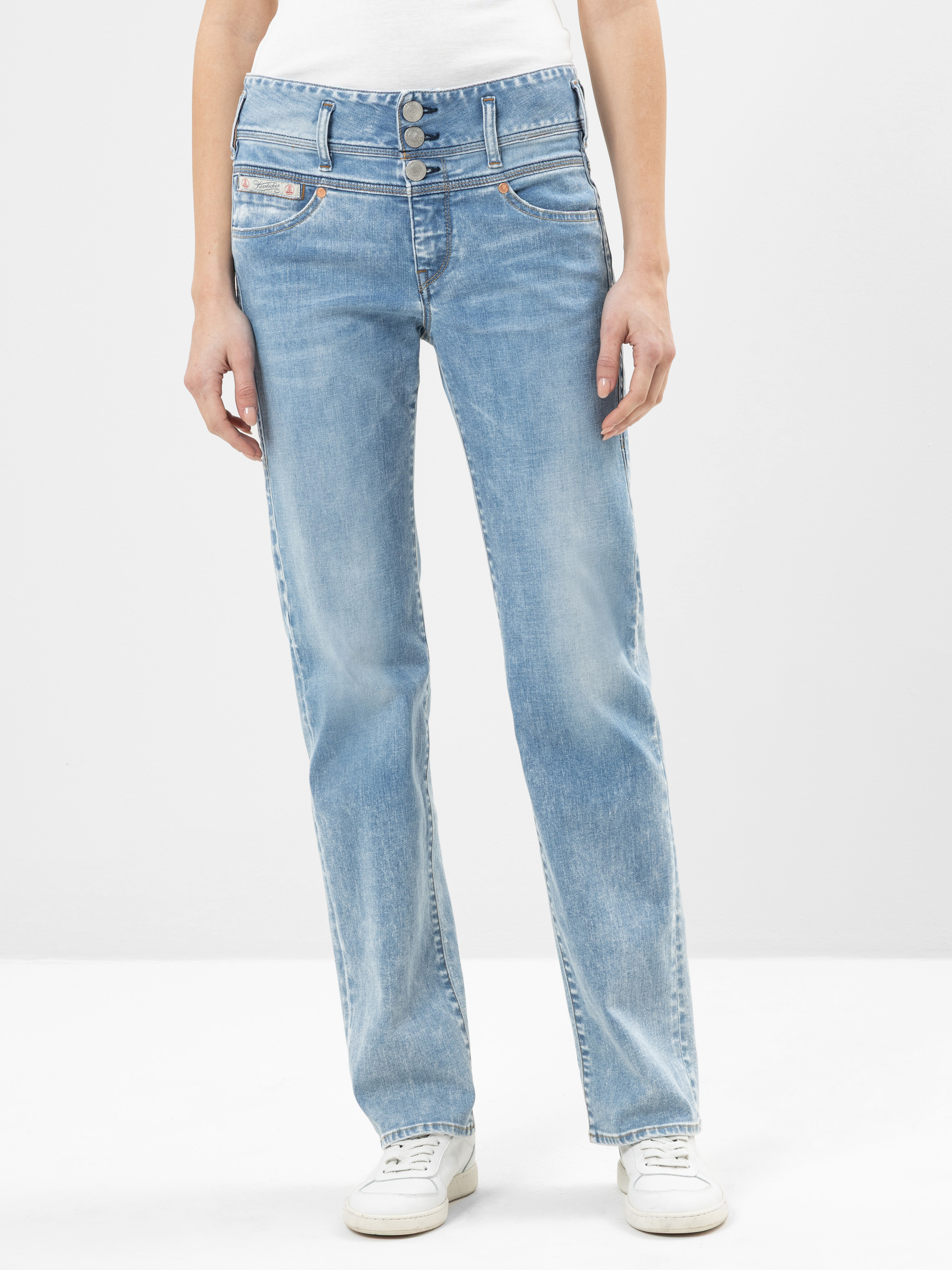 Eine Person, die hellblaue Jeans mit hoher Taille und geradem Bein mit drei Knöpfen vorne, ein weißes Oberteil und weiße Turnschuhe trägt, steht vor einem schlichten hellen Hintergrund.