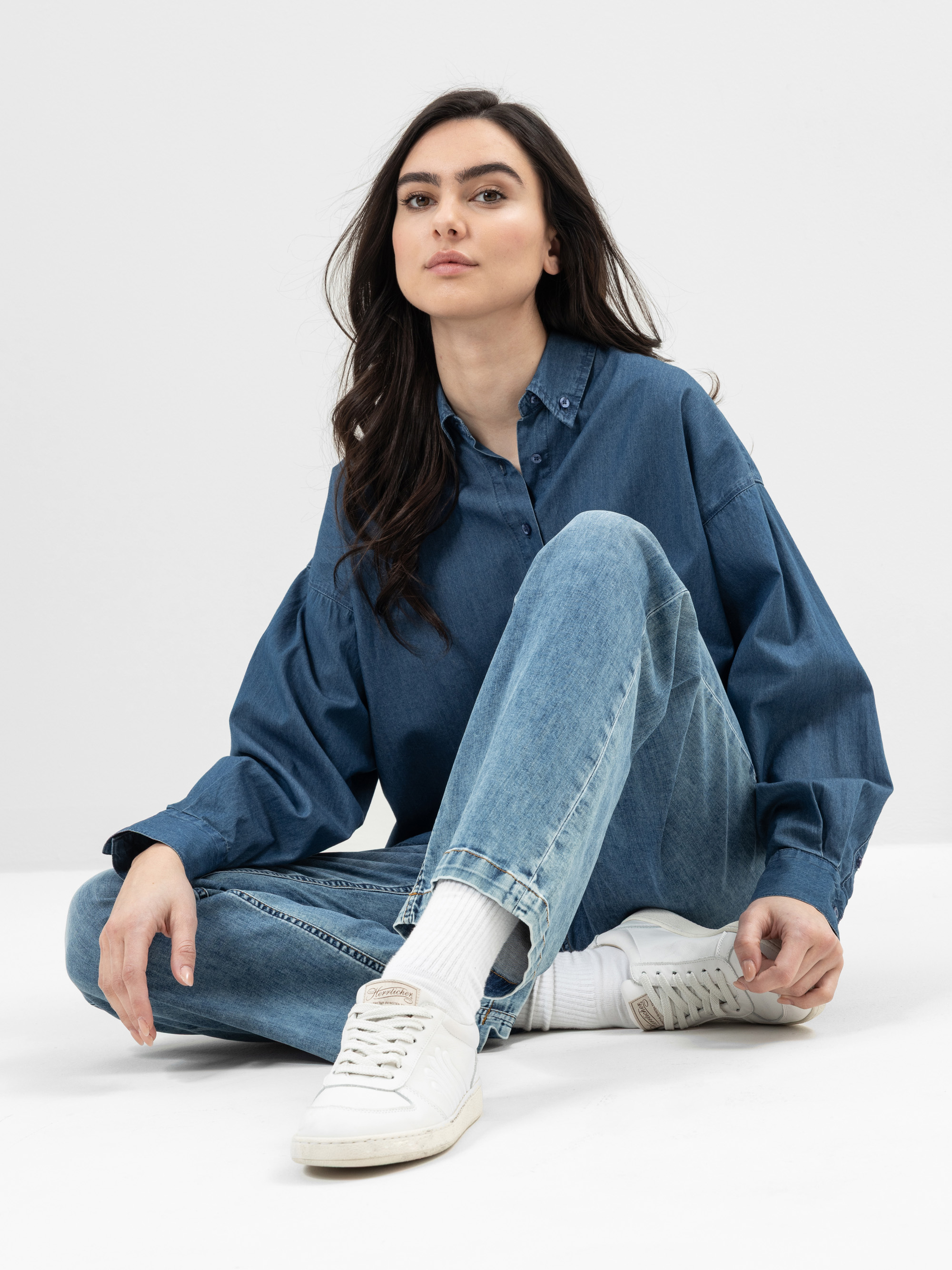 Eine Frau mit langen dunklen Haaren sitzt auf dem Boden vor einem schlichten Hintergrund, trägt ein locker sitzendes blaues Jeanshemd, blaue Jeans, weiße Socken und weiße Turnschuhe und blickt selbstbewusst in die Kamera.