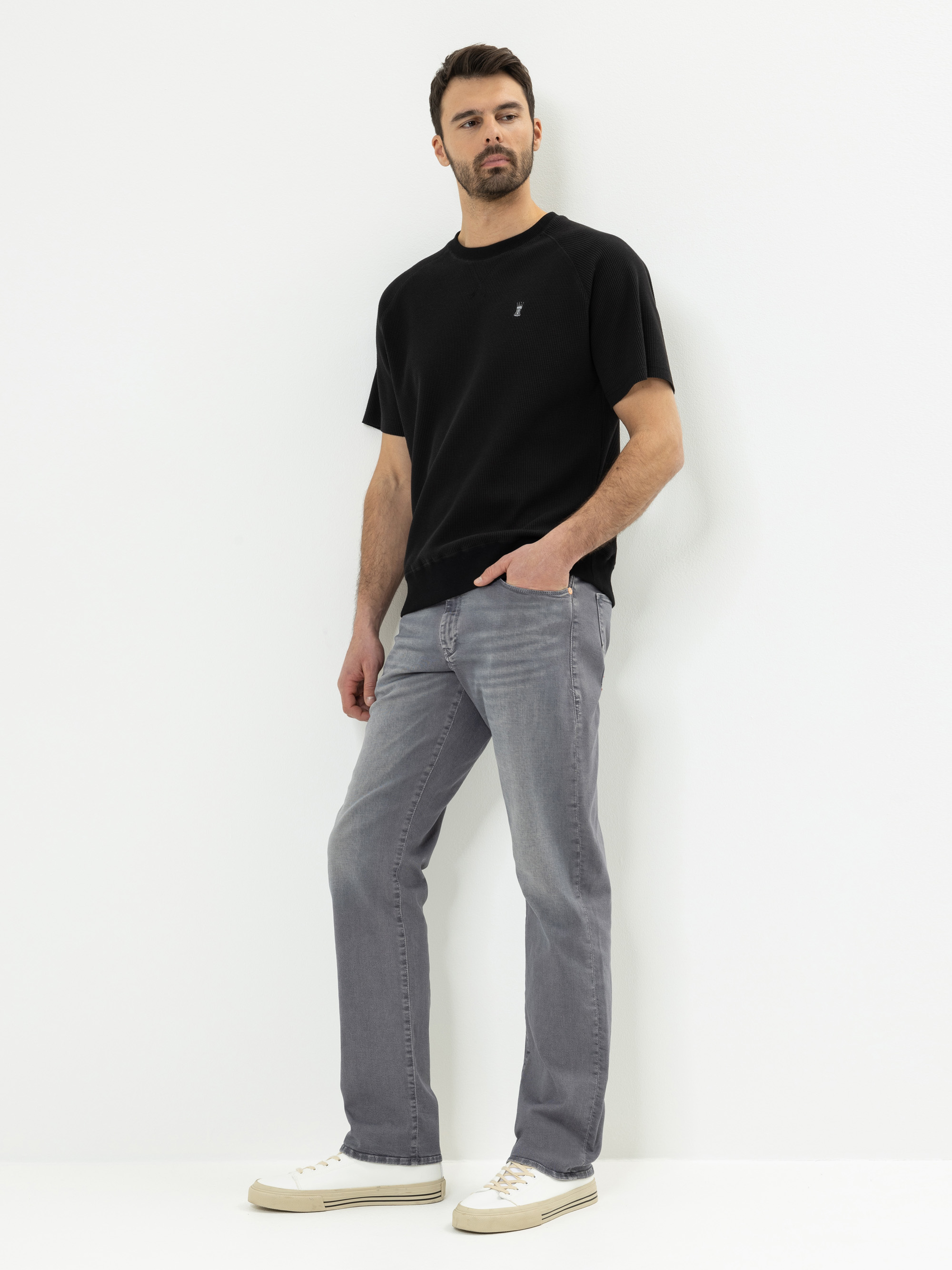 Ein Mann steht vor einem schlichten weißen Hintergrund und trägt ein schwarzes kurzärmeliges T-Shirt, hellgraue Jeans und weiße Turnschuhe. Er hat eine Hand in der Tasche und einen neutralen Gesichtsausdruck.