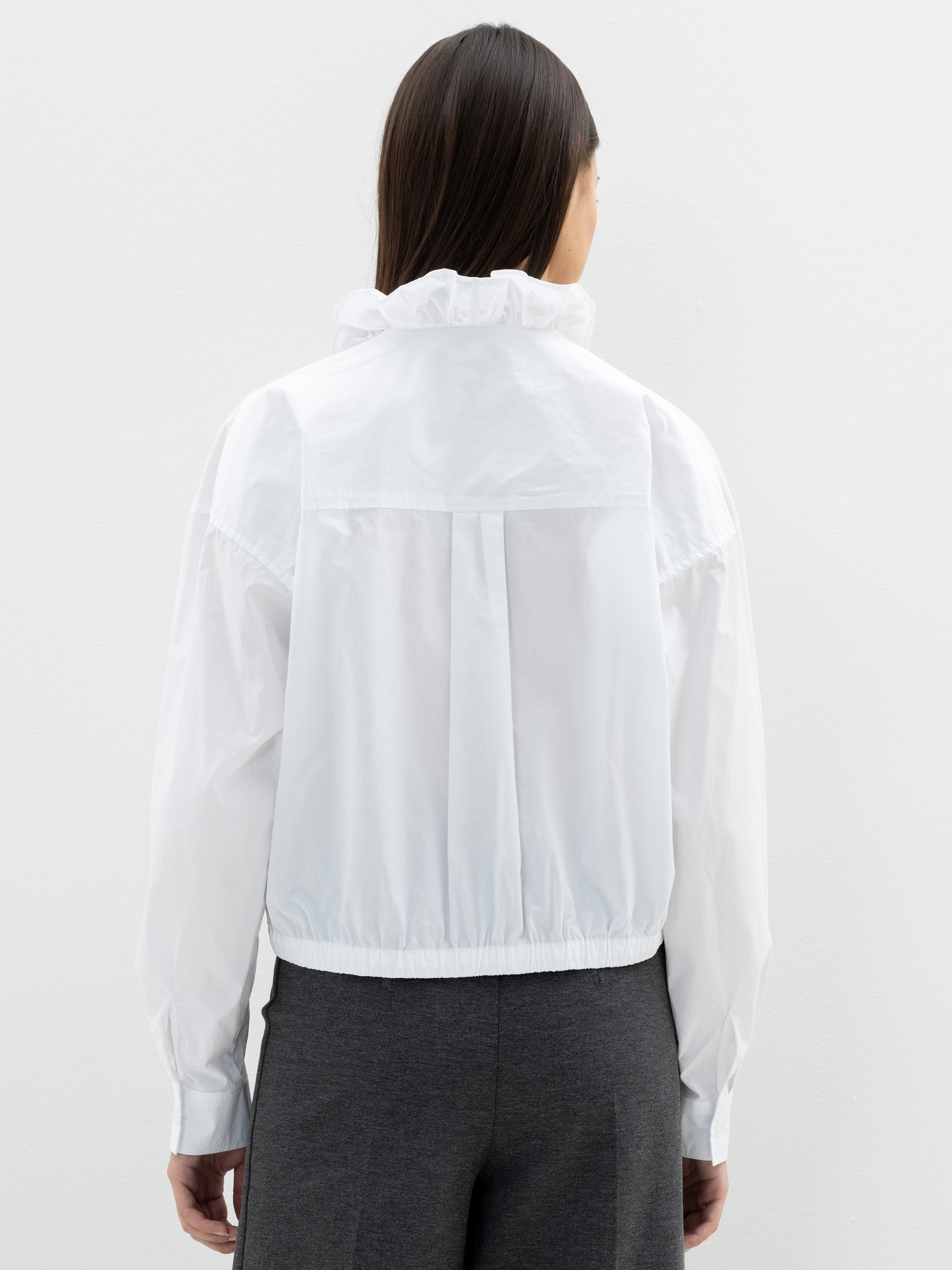 Eine Person mit langen dunklen Haaren, von hinten gesehen, trägt eine weiße, langärmelige, hochgeschlossene Bluse mit Rüschenverzierung und eine graue Hose und steht vor einem schlichten weißen Hintergrund.