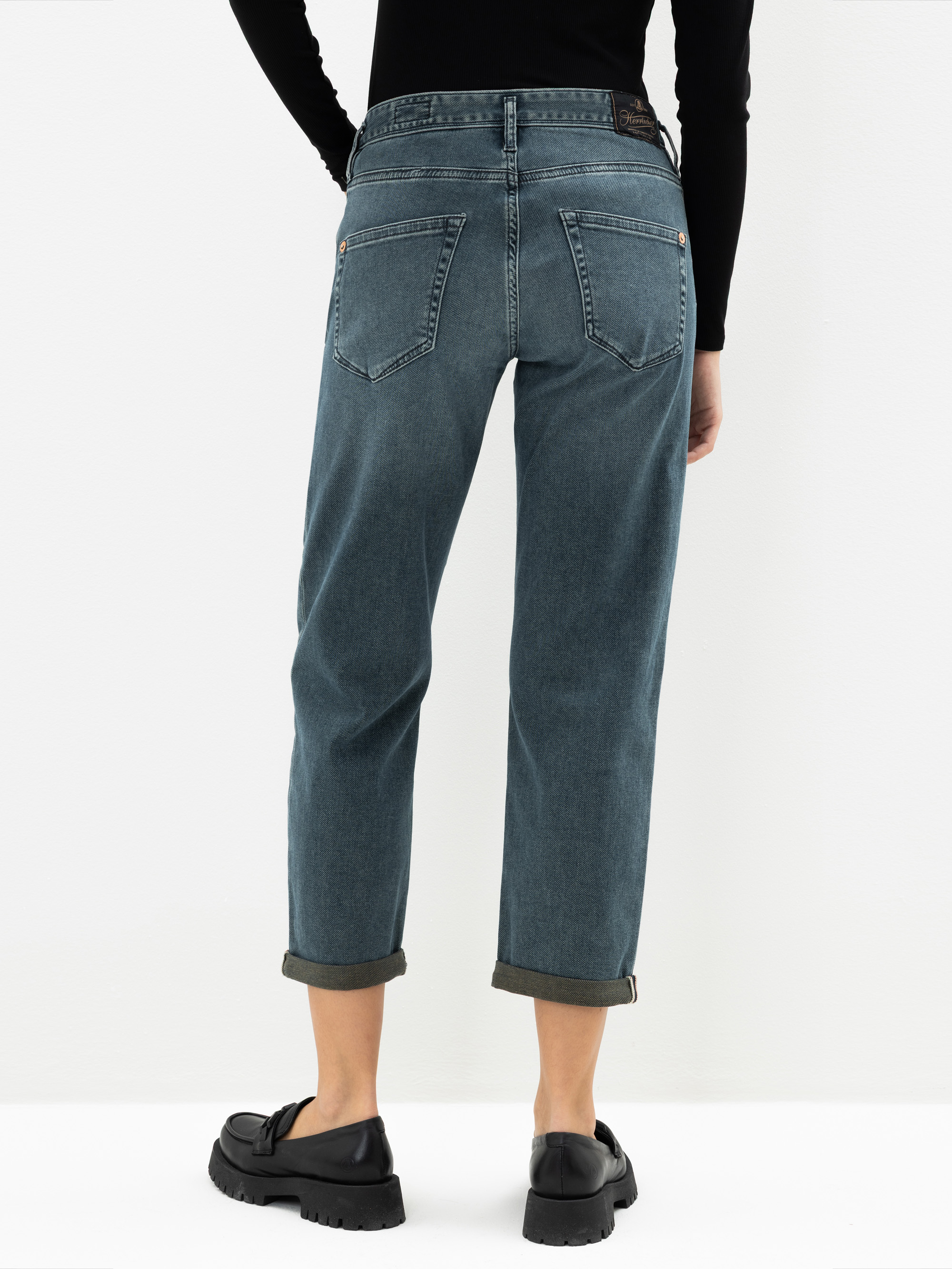 Die Person steht mit dem Gesicht zur Wand und trägt eine blaue Jeans mit Bündchen und geradem Bein, schwarze Halbschuhe und ein langärmeliges schwarzes Oberteil vor einem schlichten weißen Hintergrund. Nur die untere Hälfte des Körpers ist sichtbar.