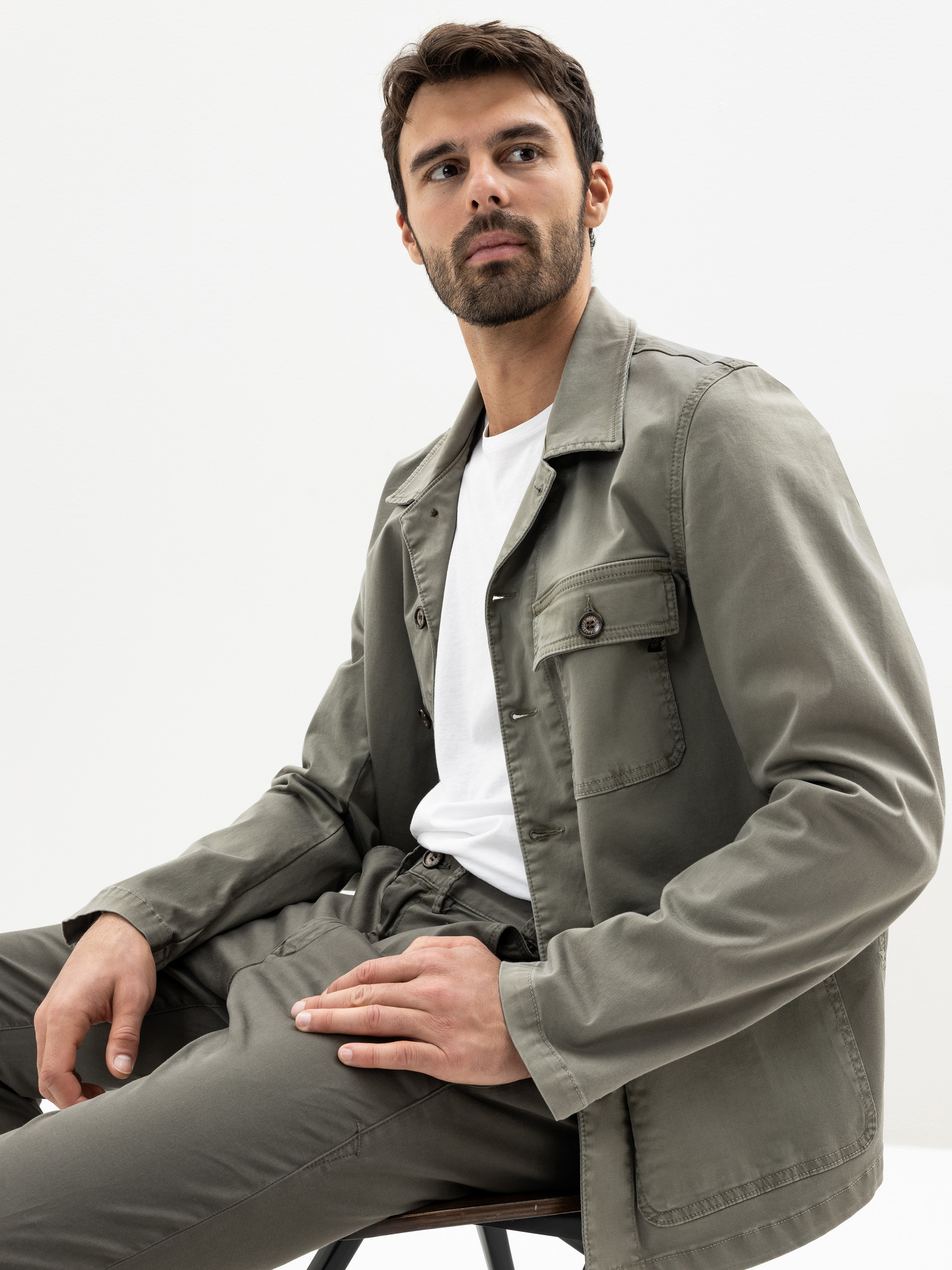 Ein Mann mit dunklem Haar und Bart sitzt auf einem Hocker und trägt eine olivgrüne Jacke und Hose mit einem weißen T-Shirt. Er blickt vor einem schlichten weißen Hintergrund leicht nach links.