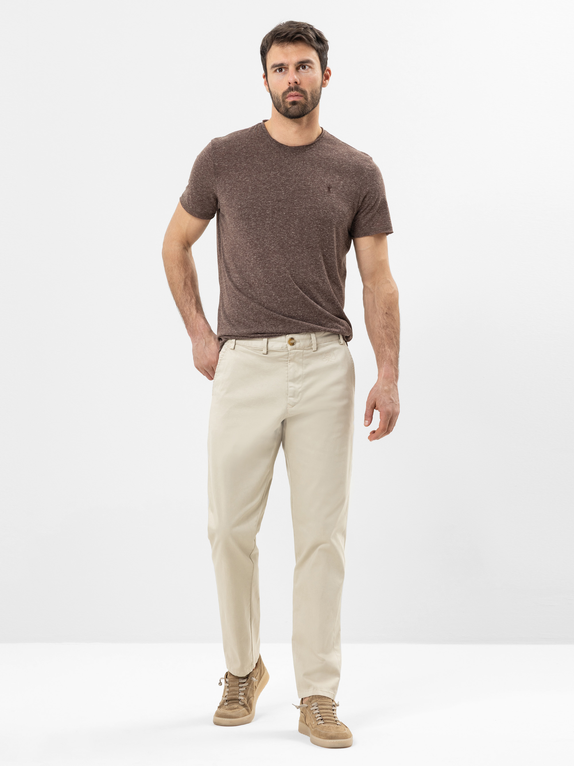 Ein Mann steht vor einem schlichten weißen Hintergrund und trägt ein kurzärmeliges braunes T-Shirt, eine hellbeige Hose und hellbraune Turnschuhe. Er hat kurzes dunkles Haar und einen Bart, eine Hand steckt in seiner Tasche.