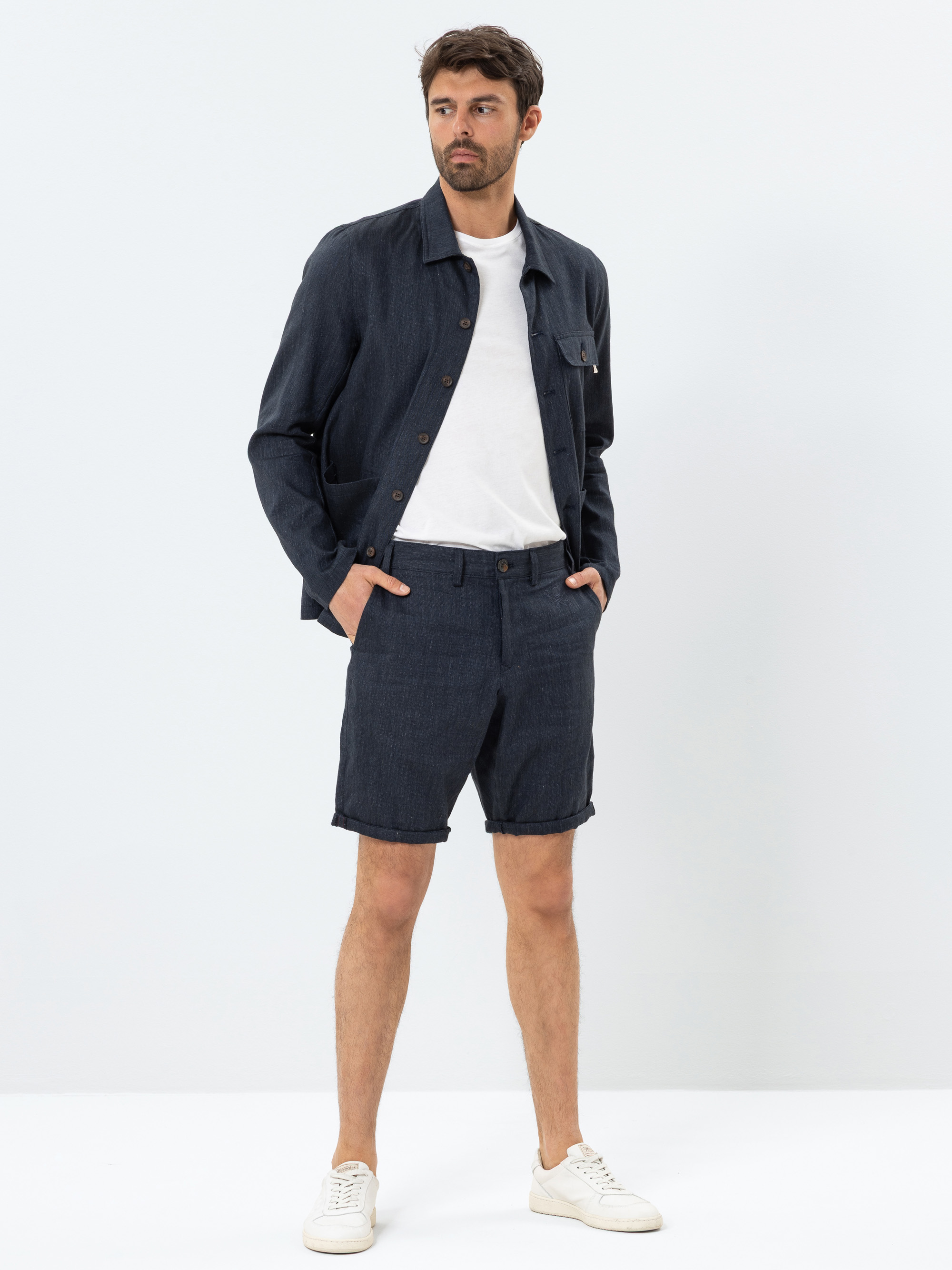 Ein Mann steht vor einem schlichten weißen Hintergrund und trägt ein marineblaues Button-up-Hemd, passende marineblaue Shorts, ein weißes T-Shirt und weiße Turnschuhe. Er hat eine Hand in der Tasche und blickt zur Seite.