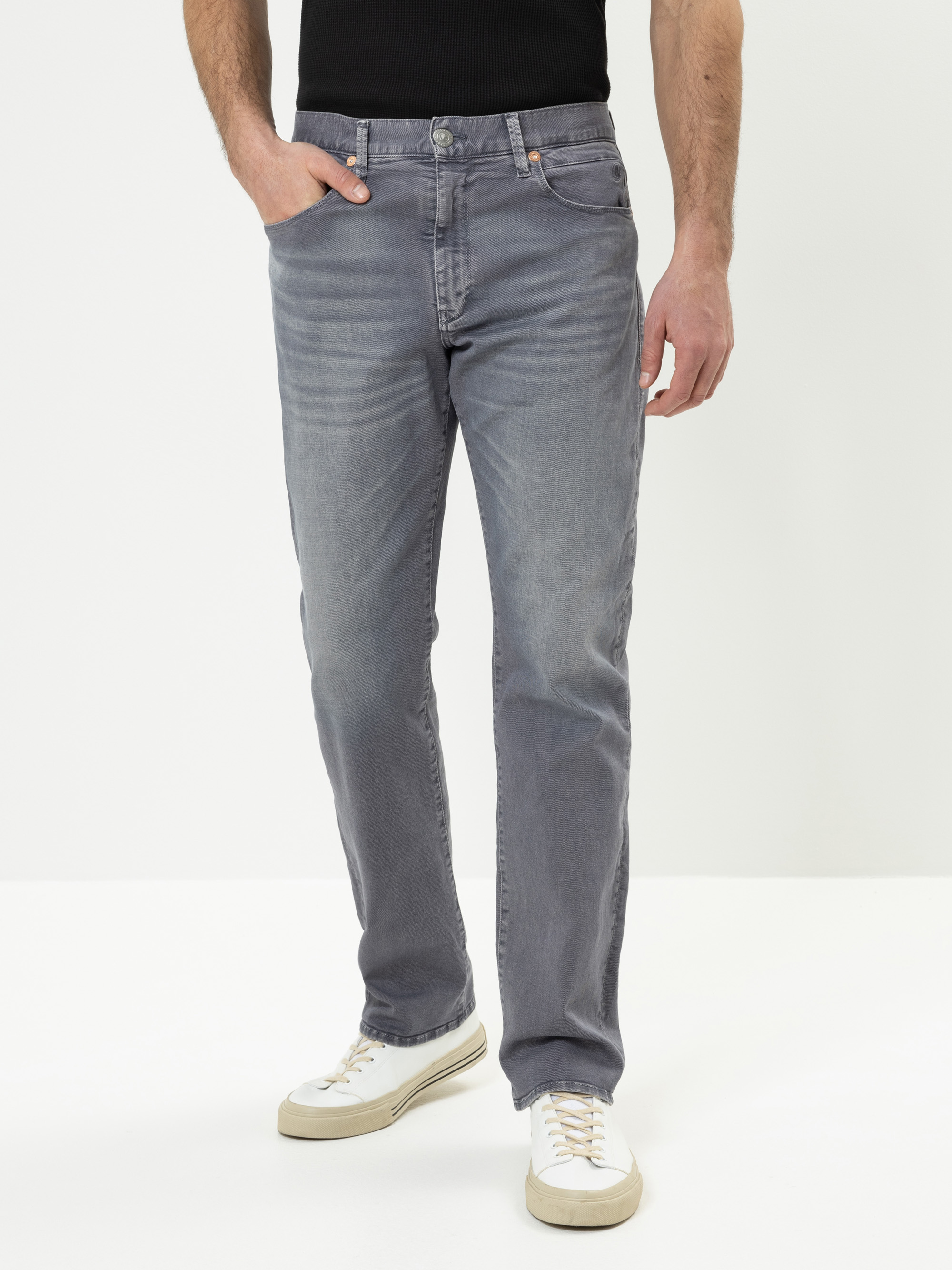 Ein Mann in verblichenen grauen Jeans, einem schwarzen Hemd und weißen Turnschuhen steht mit einer Hand in der Tasche vor einem schlichten weißen Hintergrund. Sein Gesicht ist nicht zu erkennen.