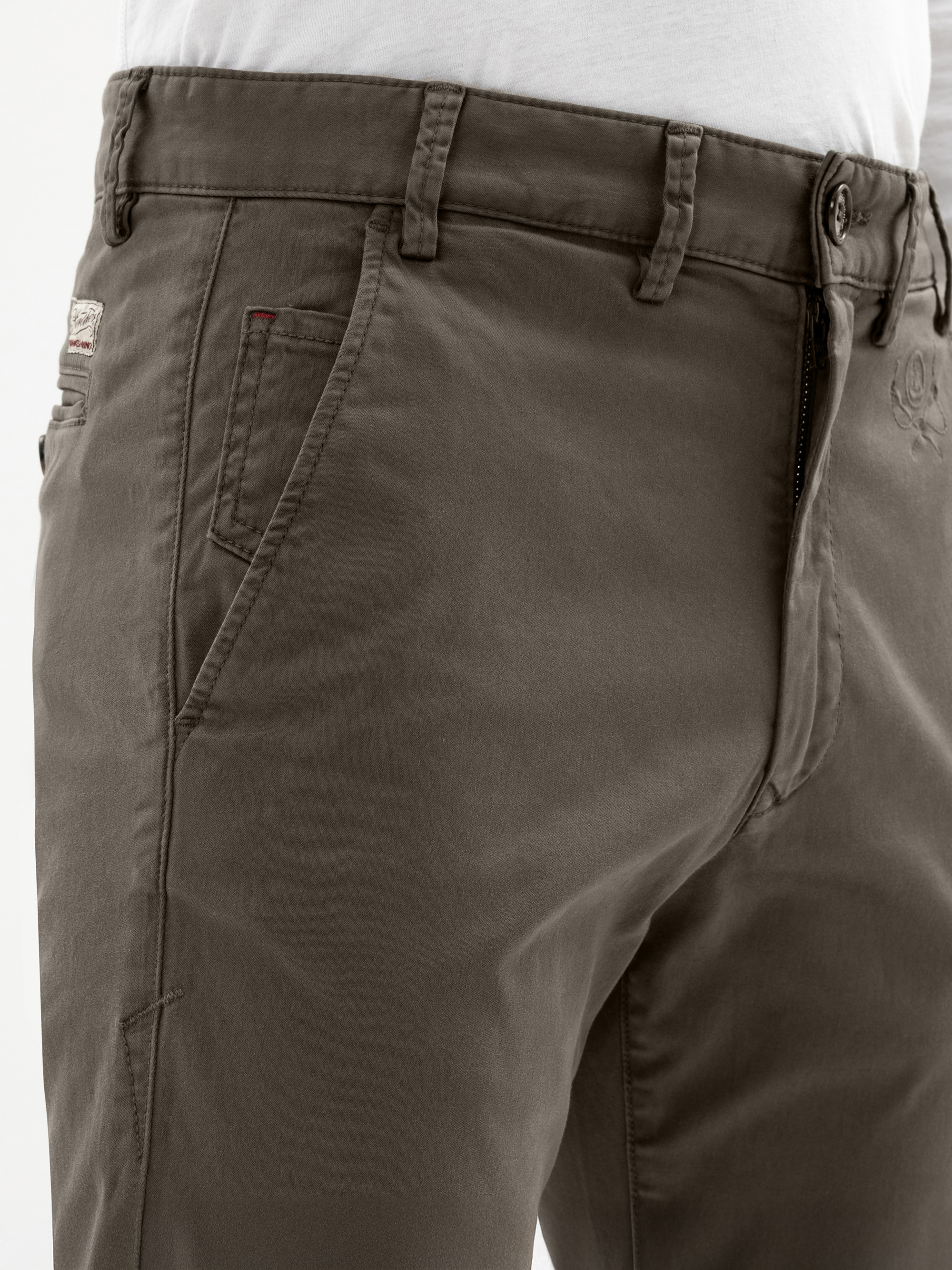 Nahaufnahme einer Person, die eine olivgrüne Hose mit teilweise geöffnetem Hosenschlitz, Gürtelschlaufen und Seitentaschen sowie ein weißes Hemd trägt, das in die Hose gesteckt wird. Die Hose weist sichtbare Nähte und ein kleines Markenetikett in de