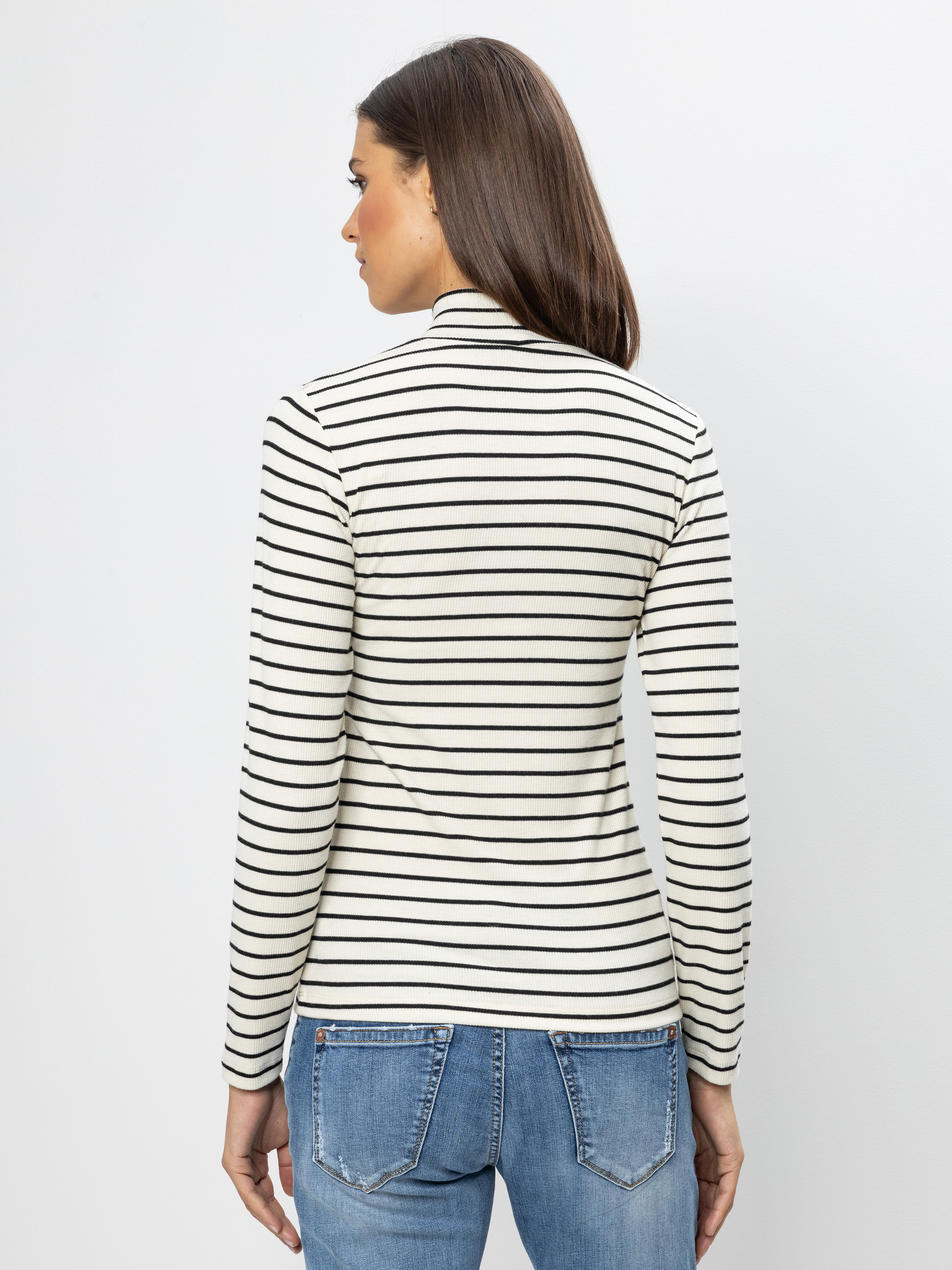 Eine Frau steht mit dem Gesicht zur Kamera, trägt ein langärmeliges, weißes Rollkragenoberteil mit schwarzen Querstreifen und blaue Jeans vor einem schlichten, hellen Hintergrund.