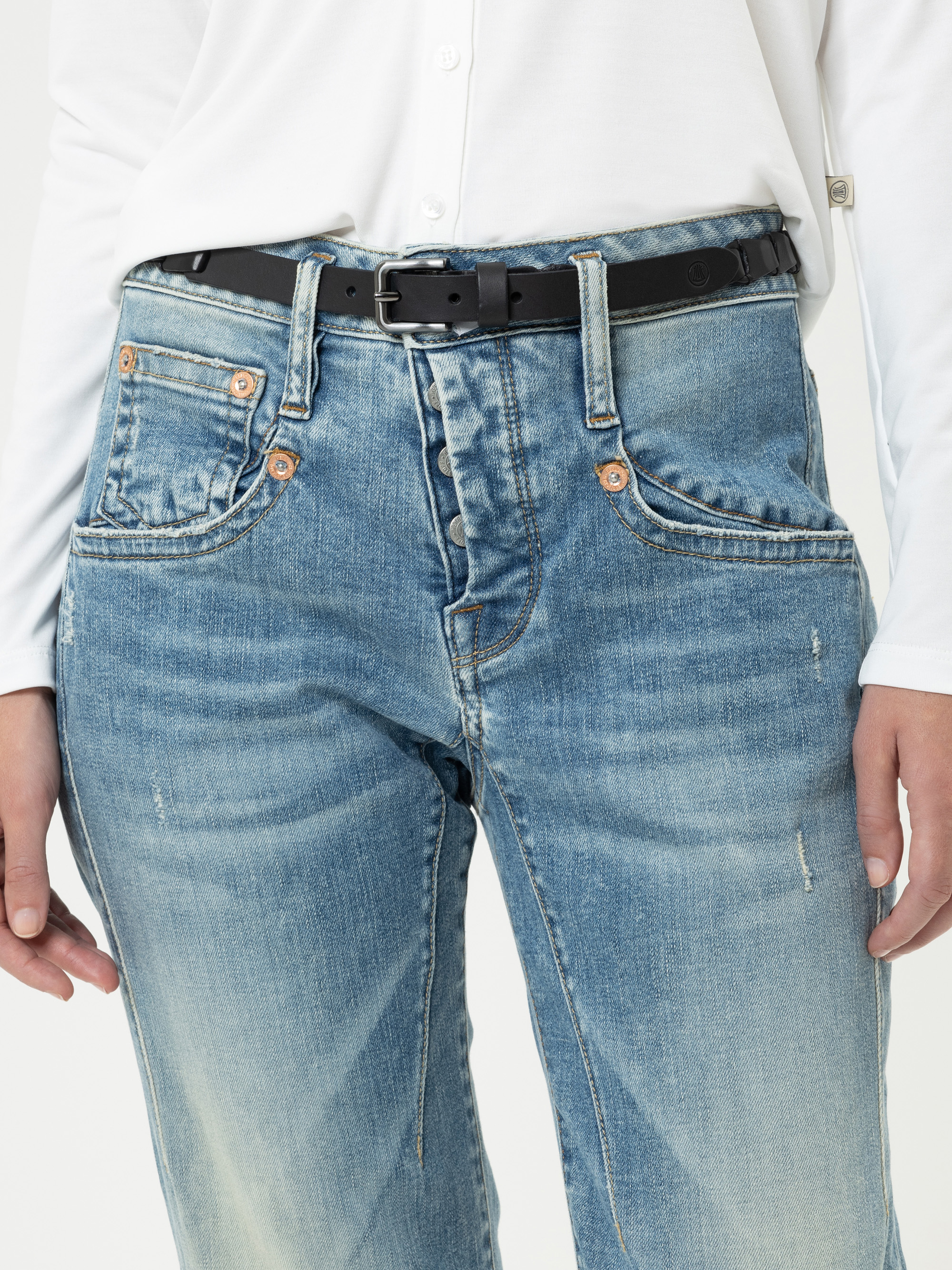 Eine Person, die ein weißes langärmeliges Hemd, eine hellblaue Jeans mit sichtbaren Nähten und kupferfarbenen Knöpfen sowie einen schwarzen Gürtel trägt. Das Foto ist von den Schultern bis zur Mitte des Oberschenkels abgeschnitten.