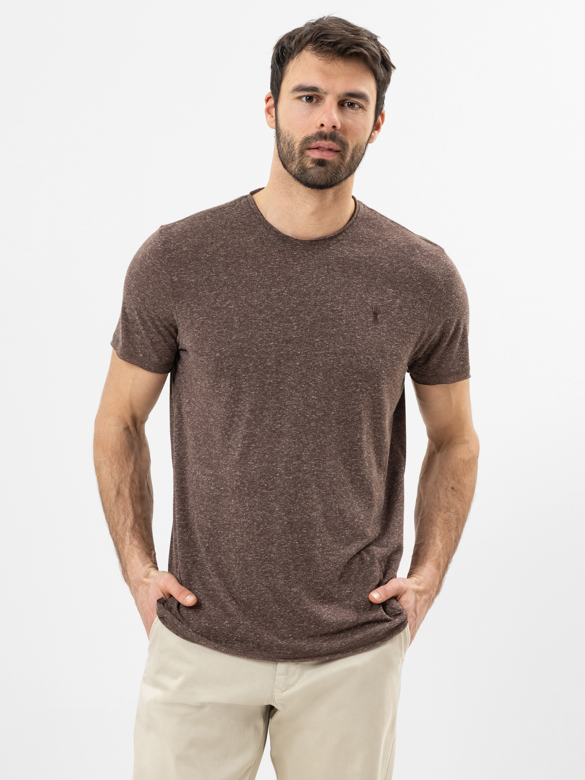 Ein Mann mit kurzen dunklen Haaren und einem Bart trägt ein braunes kurzärmeliges T-Shirt und eine beigefarbene Hose. Er steht vor einem schlichten weißen Hintergrund, hat die Hände in den Taschen und blickt in die Kamera.