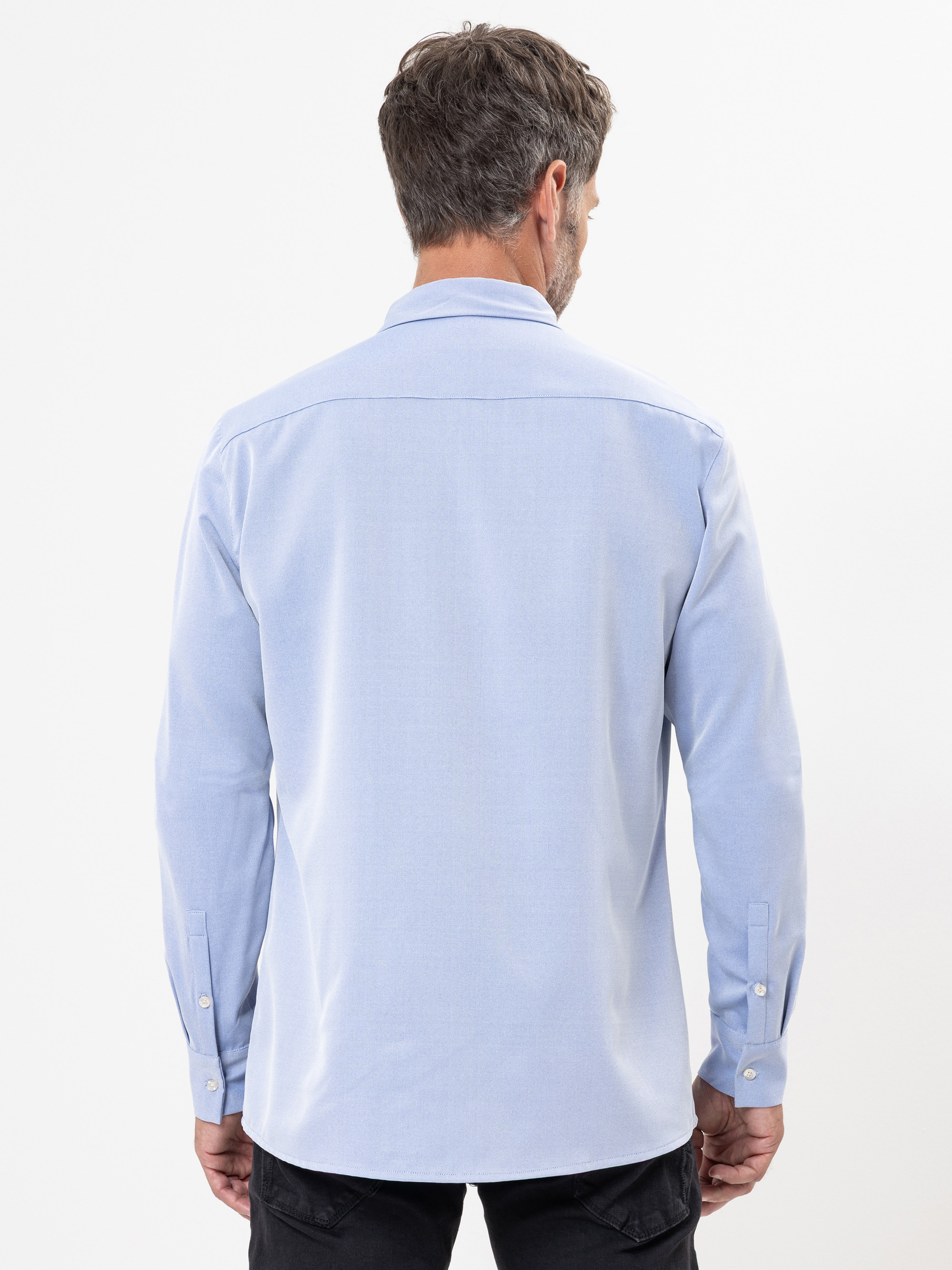 Ein Mann mit kurzen braunen Haaren ist von hinten zu sehen. Er trägt ein hellblaues, langärmeliges Hemd mit Knopfleiste und eine schwarze Hose vor einem schlichten weißen Hintergrund.