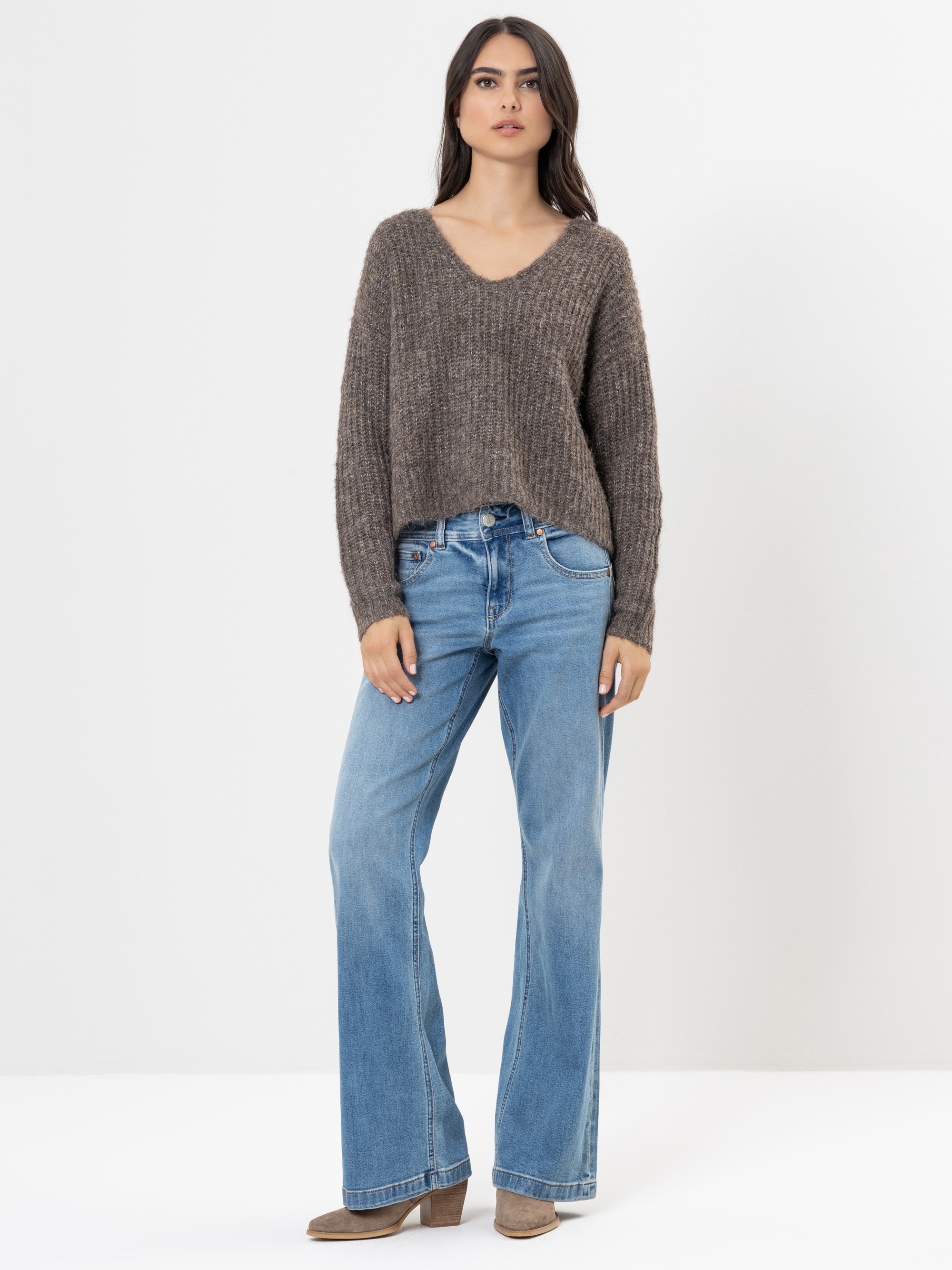 Eine Frau steht vor einem schlichten weißen Hintergrund und trägt einen braunen, langärmeligen Strickpullover, hellblaue, weit geschnittene Jeans und braune Stiefel. Sie hat langes dunkles Haar und einen neutralen Gesichtsausdruck.