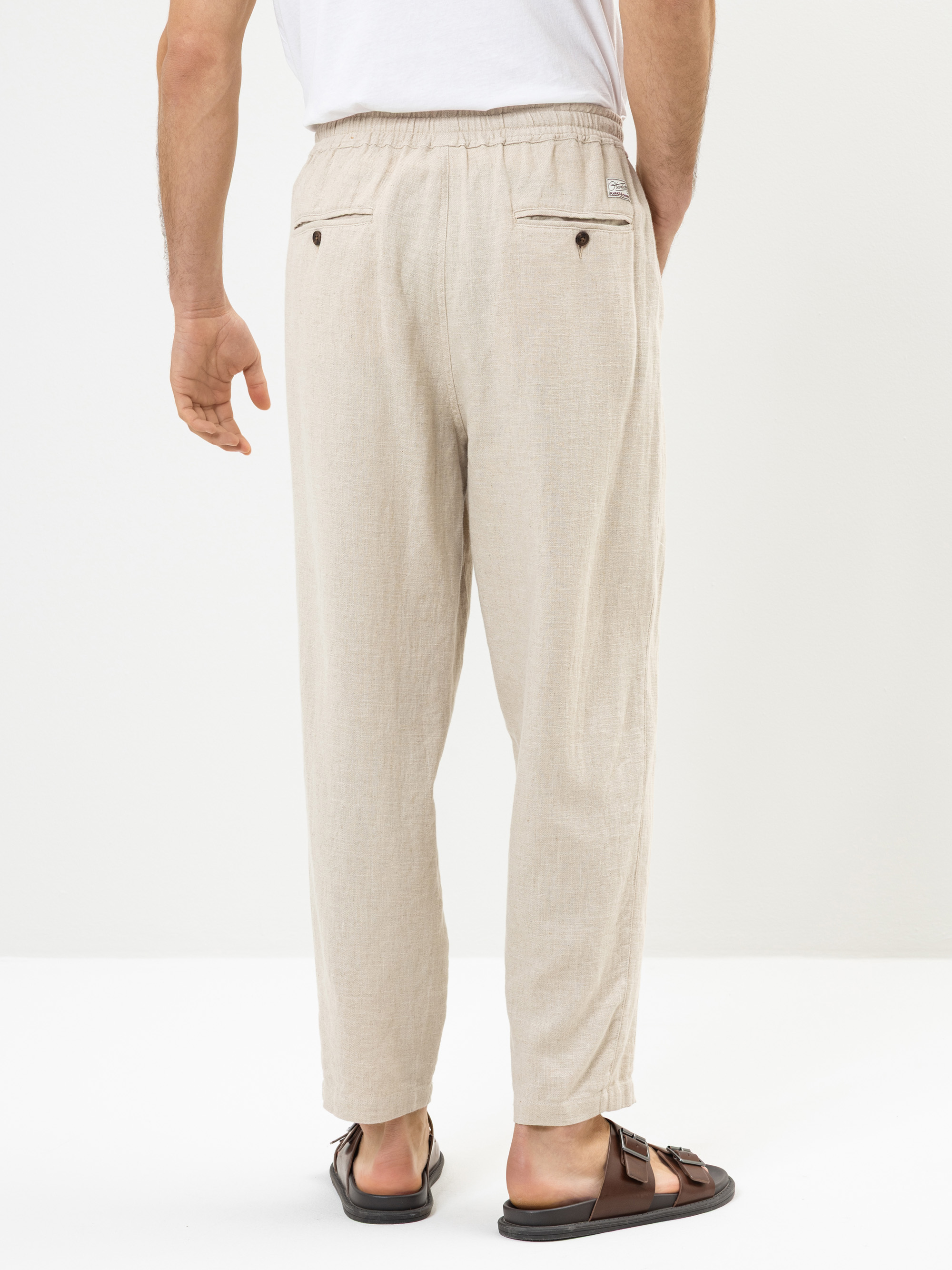 Eine Person, die eine hellbeige, locker sitzende Hose mit elastischem Bund und geknöpften Gesäßtaschen, ein weißes T-Shirt und braune Sandalen trägt, steht vor einem schlichten weißen Hintergrund.