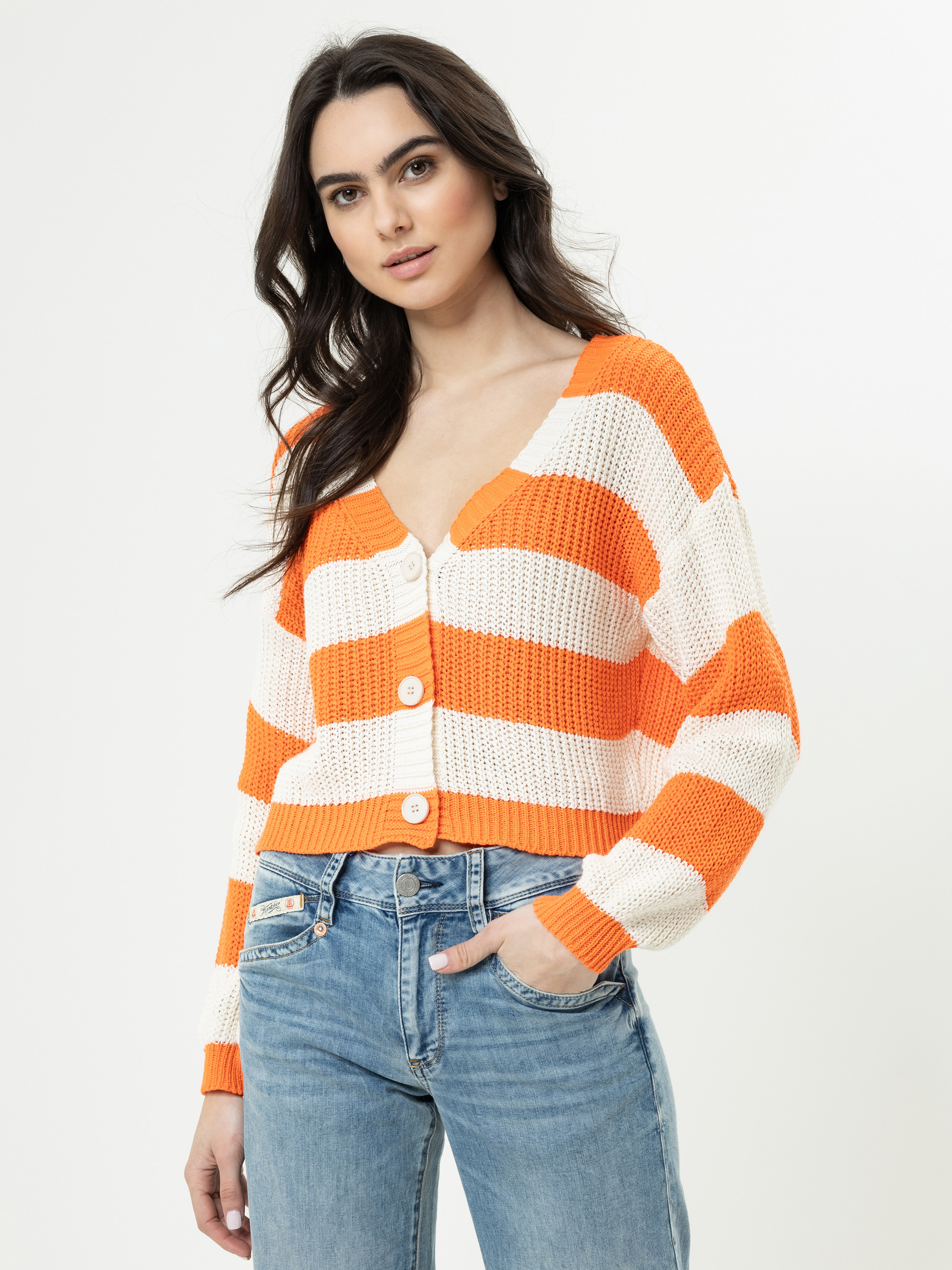 Eine Frau, die eine orange-weiß gestreifte Strickjacke mit großen Knöpfen und blaue Jeans trägt, steht vor einem schlichten weißen Hintergrund.