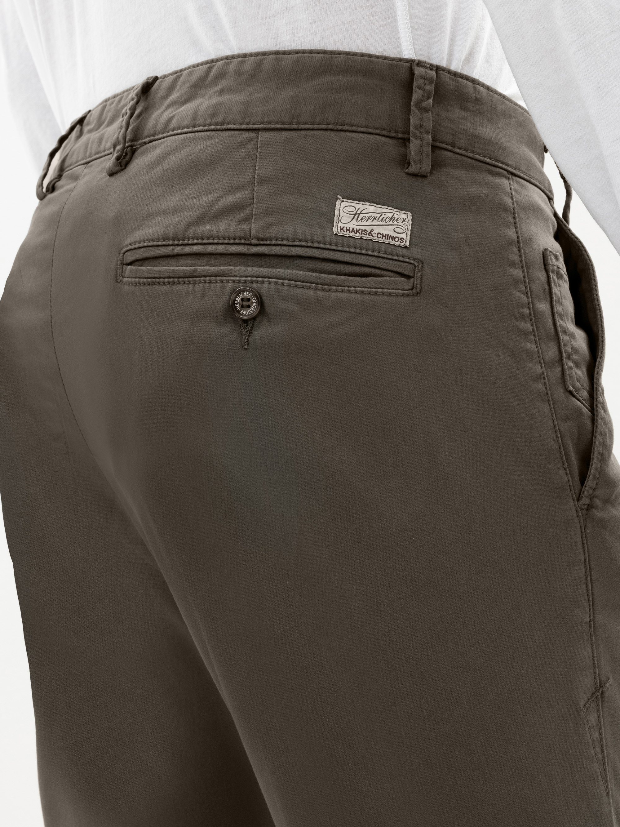 Nahaufnahme einer Person, die eine dunkelbraune Hose mit einer Gesäßtasche mit einem Knopf und einem aufgenähten Etikett darüber trägt, dazu ein weißes Hemd.