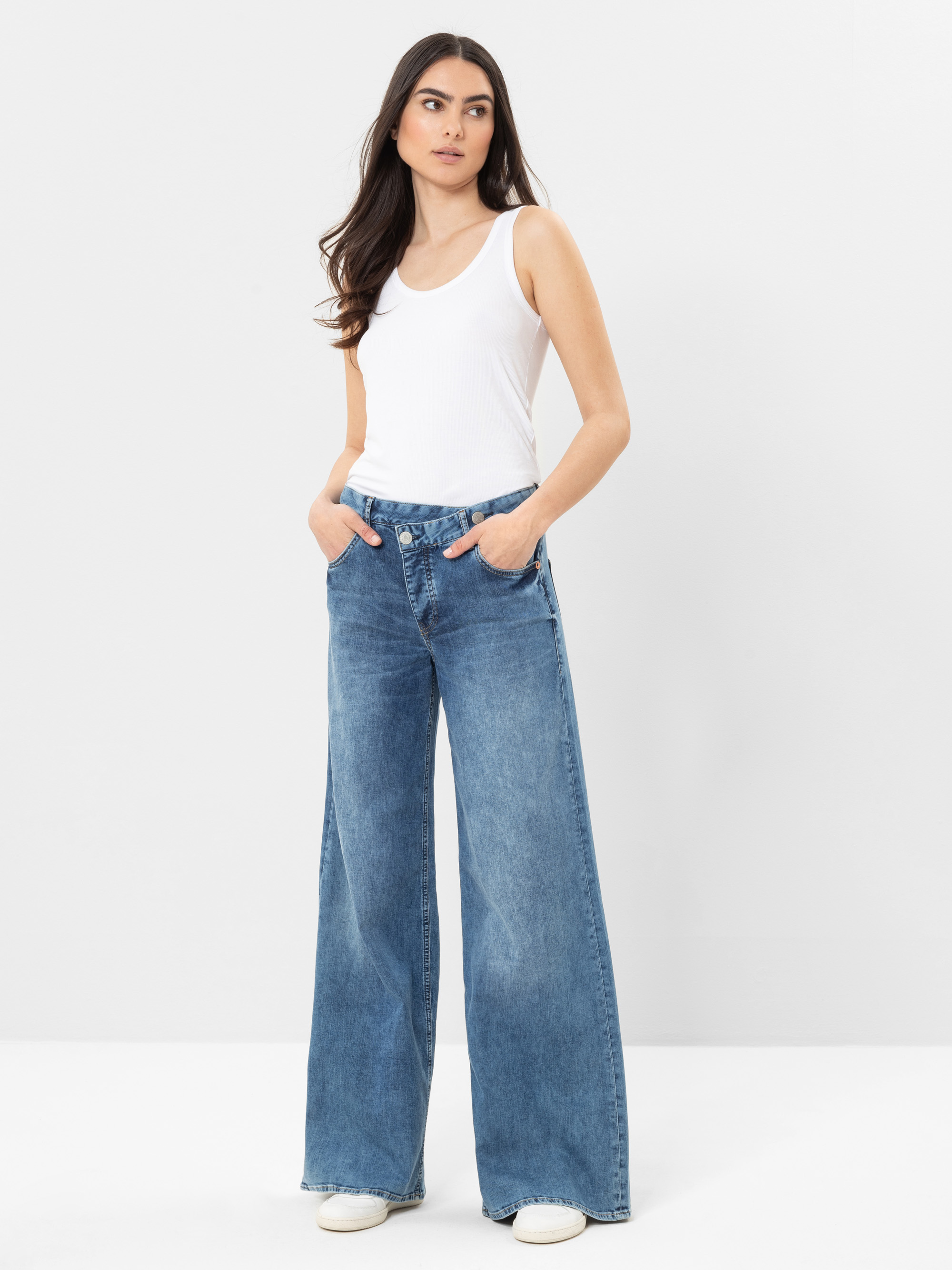 Eine Frau steht vor einem schlichten weißen Hintergrund und trägt ein weißes ärmelloses Tank-Top, hochgeschnittene Jeans mit weitem Bein und weiße Turnschuhe. Sie hat langes dunkles Haar und ihre Hände stecken in den Taschen.
