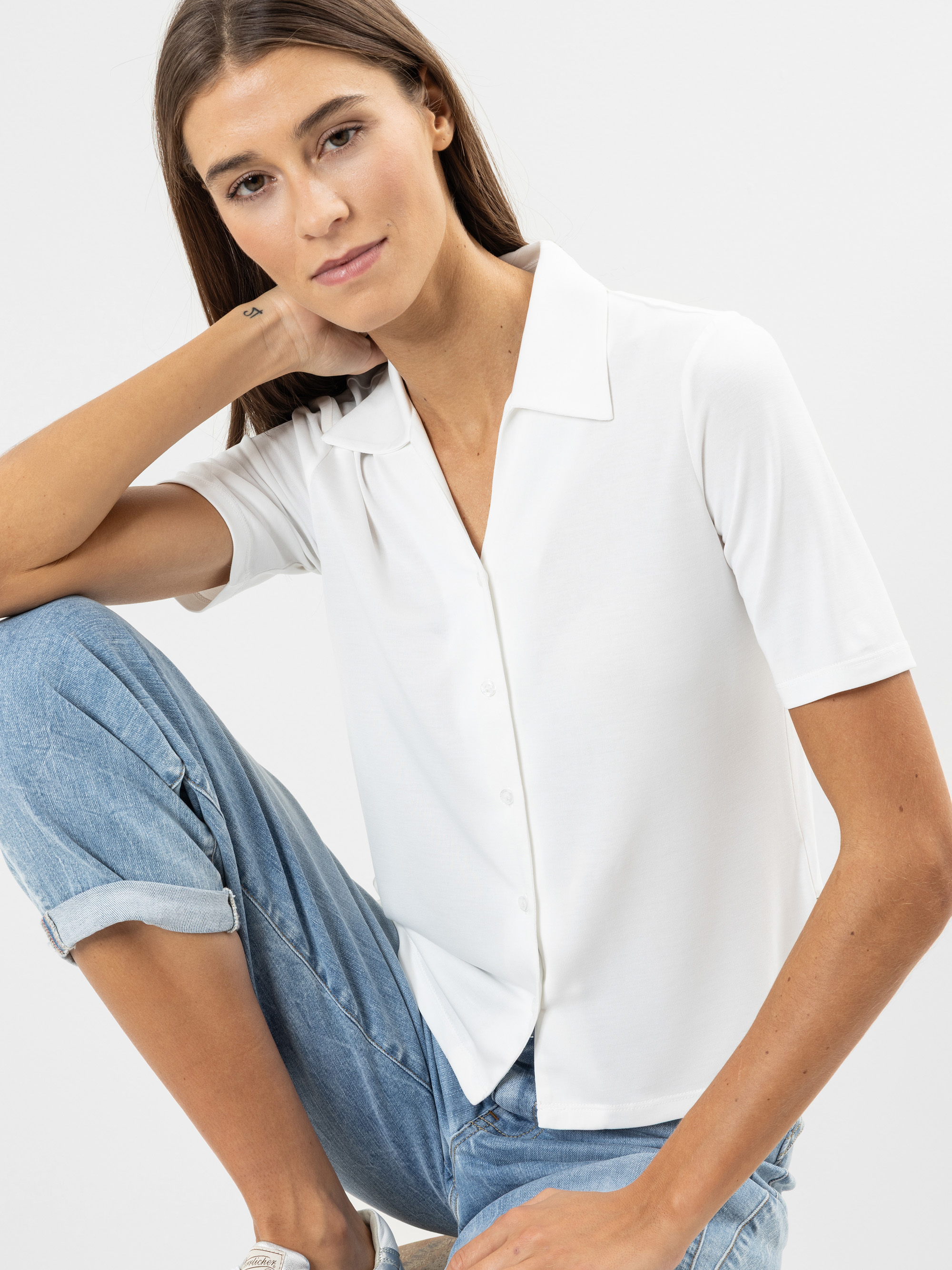 Eine Frau mit glattem braunem Haar, die ein kurzärmeliges weißes Button-up-Hemd und hellblaue, hochgekrempelte Jeans trägt, sitzt mit einem hochgezogenen Knie und blickt in die Kamera vor einem schlichten hellen Hintergrund.