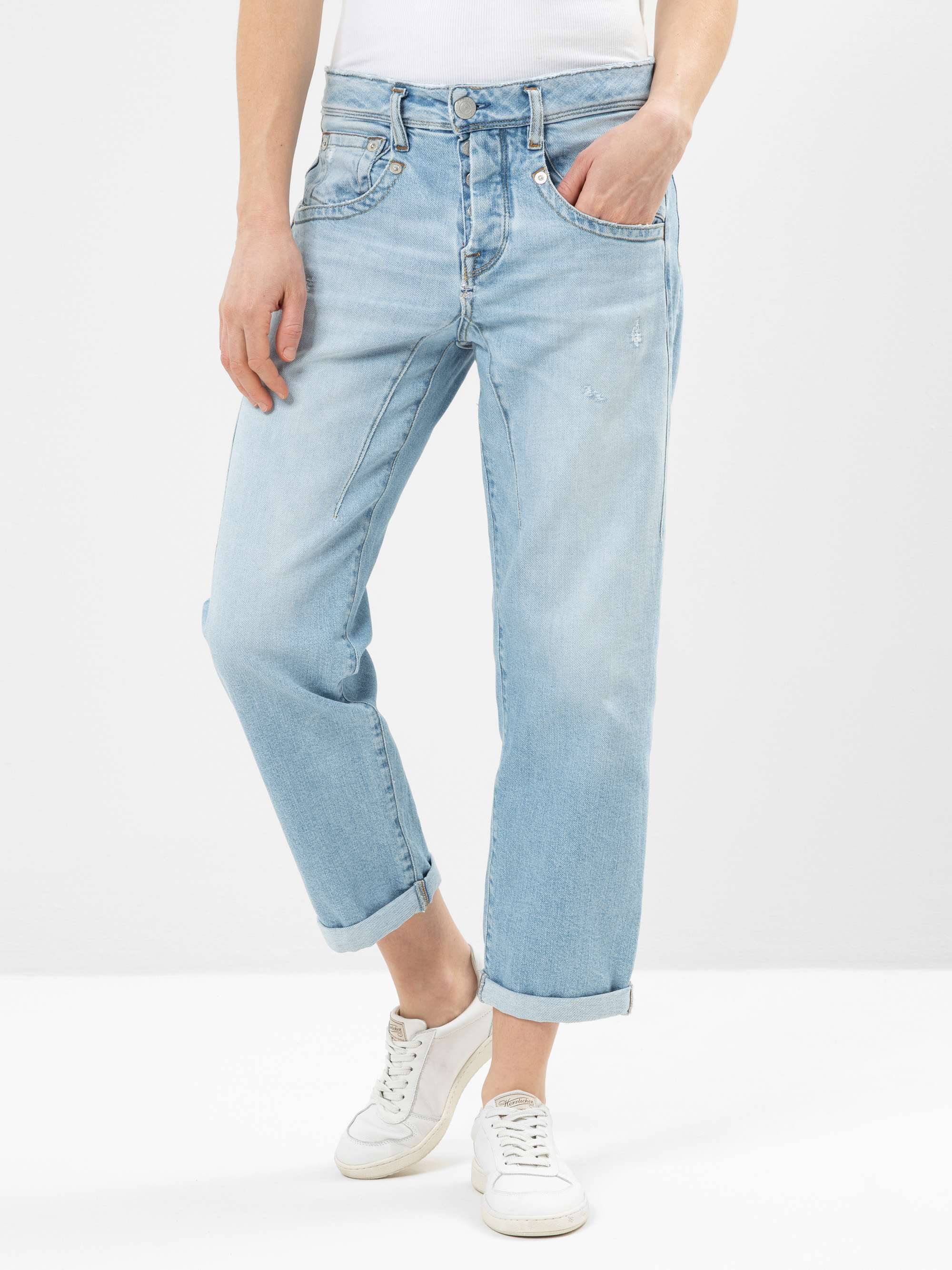 Eine Person, die hellblaue, leicht abgenutzte, locker sitzende Jeans mit aufgerollten Manschetten, ein weißes Oberteil und weiße Turnschuhe trägt und vor einem schlichten weißen Hintergrund steht.