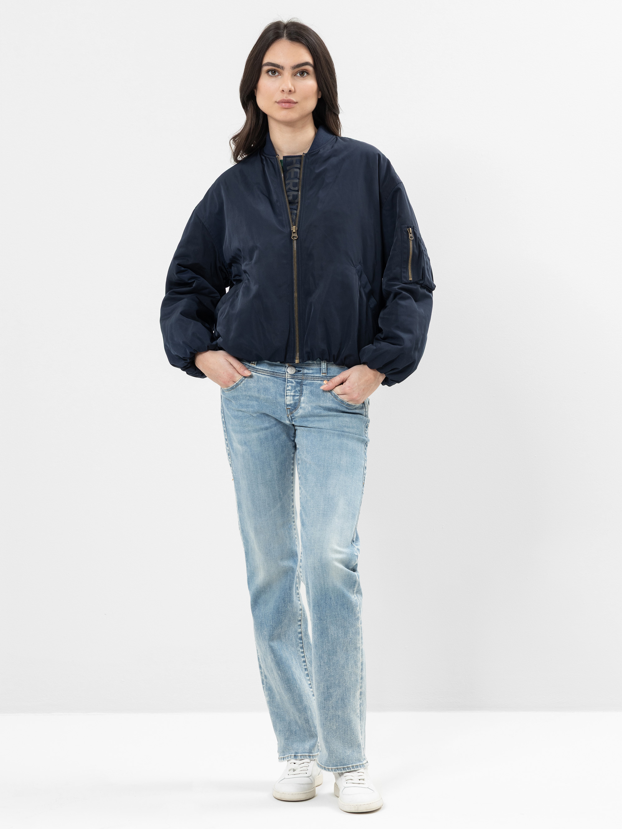 Eine Frau steht vor einem schlichten weißen Hintergrund und trägt eine dunkelblaue Bomberjacke, hellblaue Jeans und weiße Turnschuhe. Sie hat langes dunkles Haar und einen neutralen Gesichtsausdruck, die Hände stecken in den Jackentaschen.